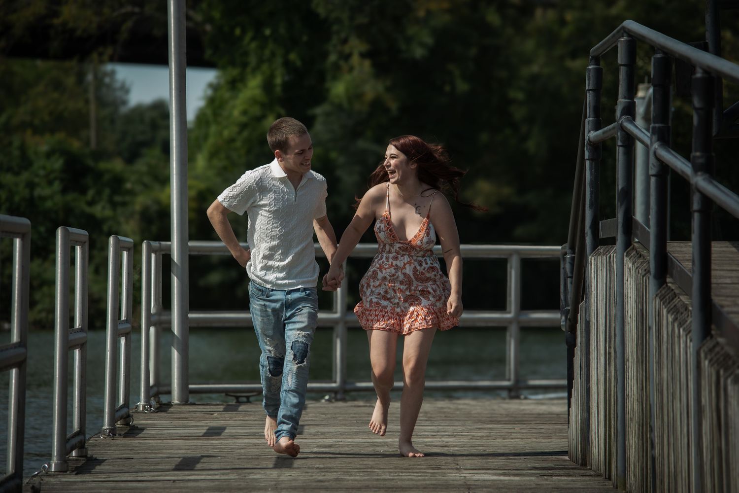 Couple walking hand-in-hand on a sunny wooden pier near a lake, woman in floral dress, man in jeans.