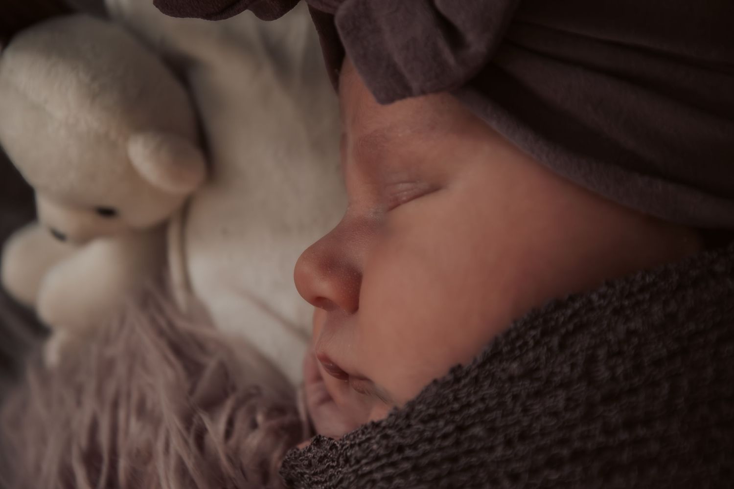 Sleeping newborn baby wrapped in gray knit blanket with white teddy bear, wearing a soft hat in dim light.