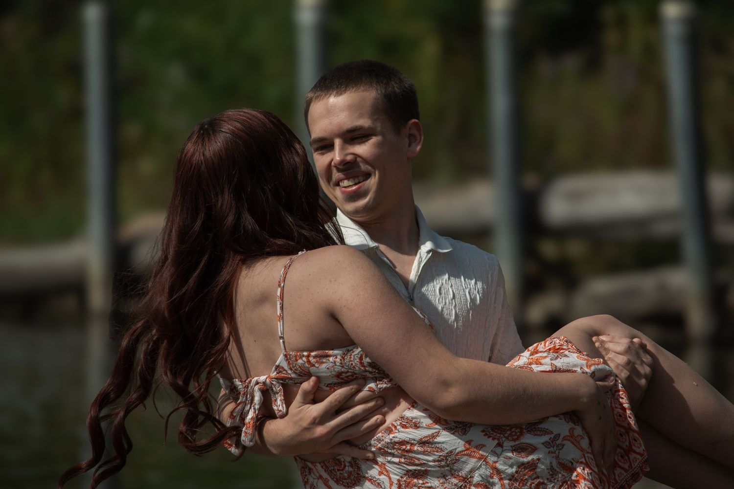 A couple embraces and smiles near a waterfront dock on a sunny day.