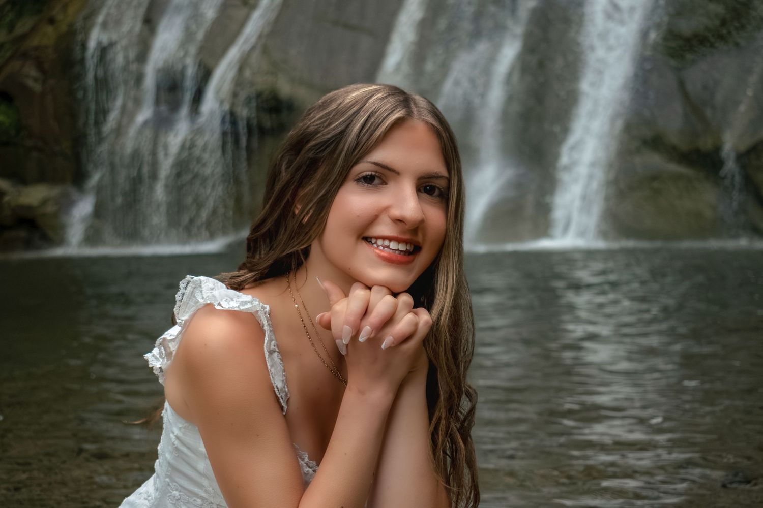 Smiling young woman in white dress posing near a waterfall with wet hair and hands clasped under chin.