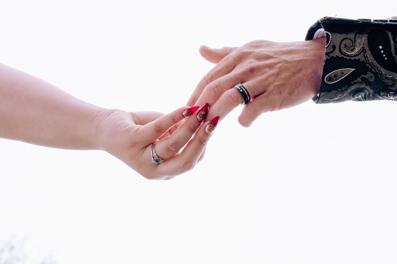 Two hands touching, one wearing a diamond ring and red nails, the other with a black ring and ornate sleeve.