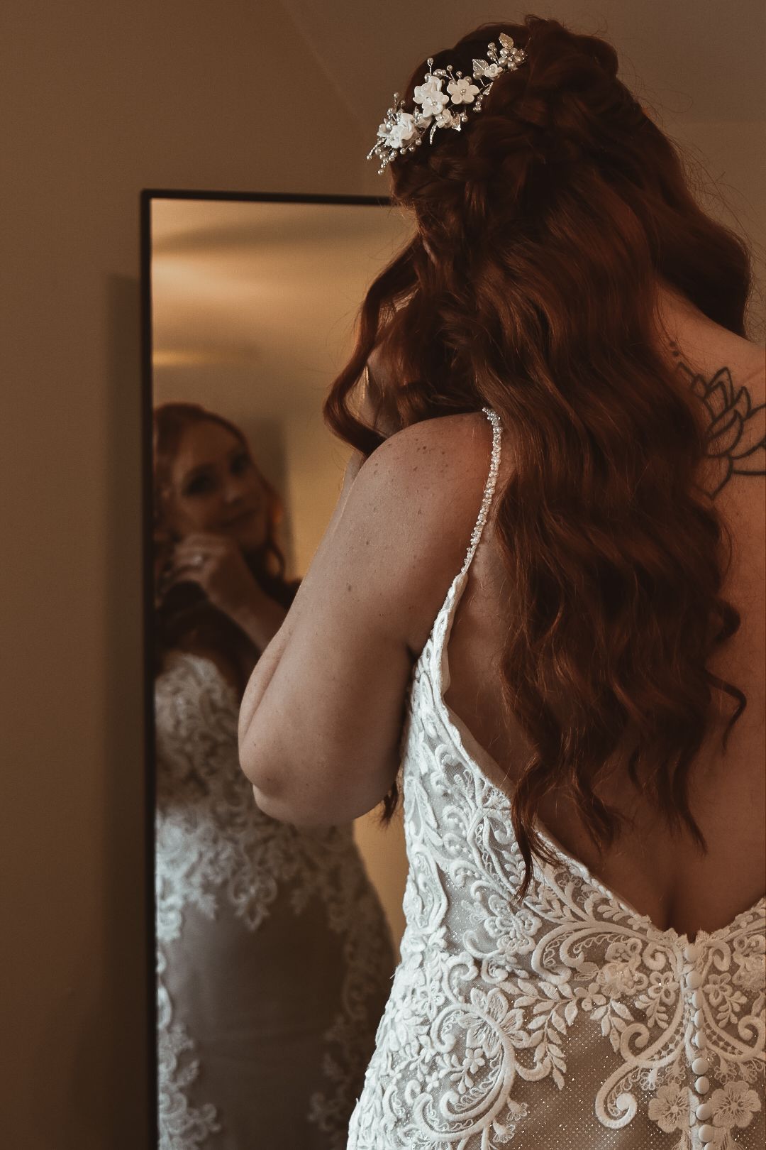 Bride in lace wedding gown with floral hair accessory views reflection in mirror, showcasing open back and long curly red hair.