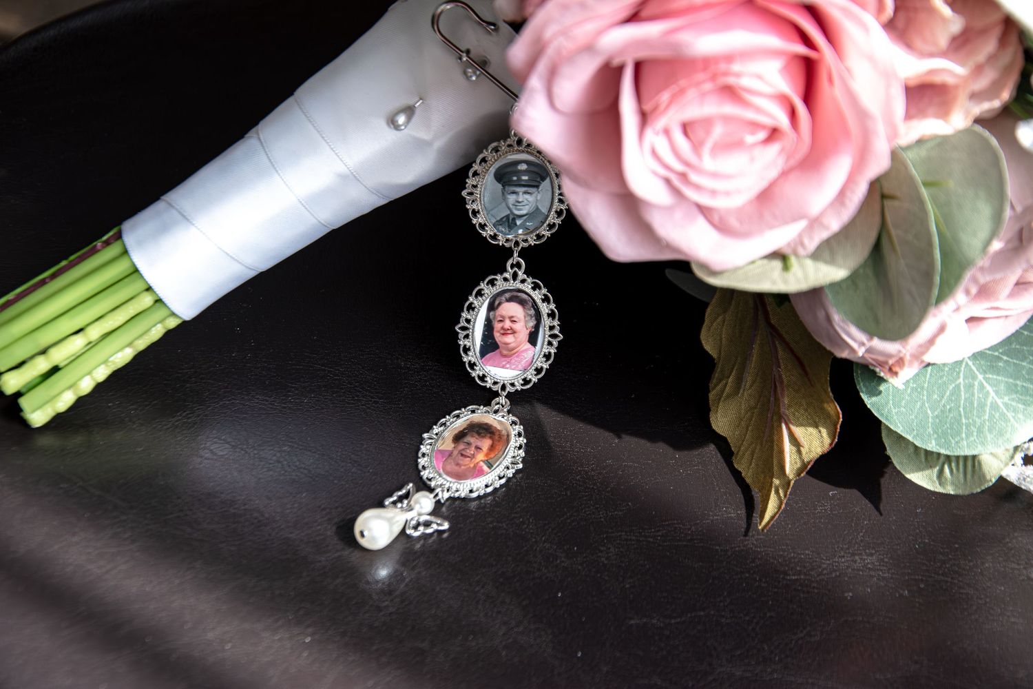 A bridal bouquet charm featuring memorial photo pendants rests beside pink roses on a dark surface.