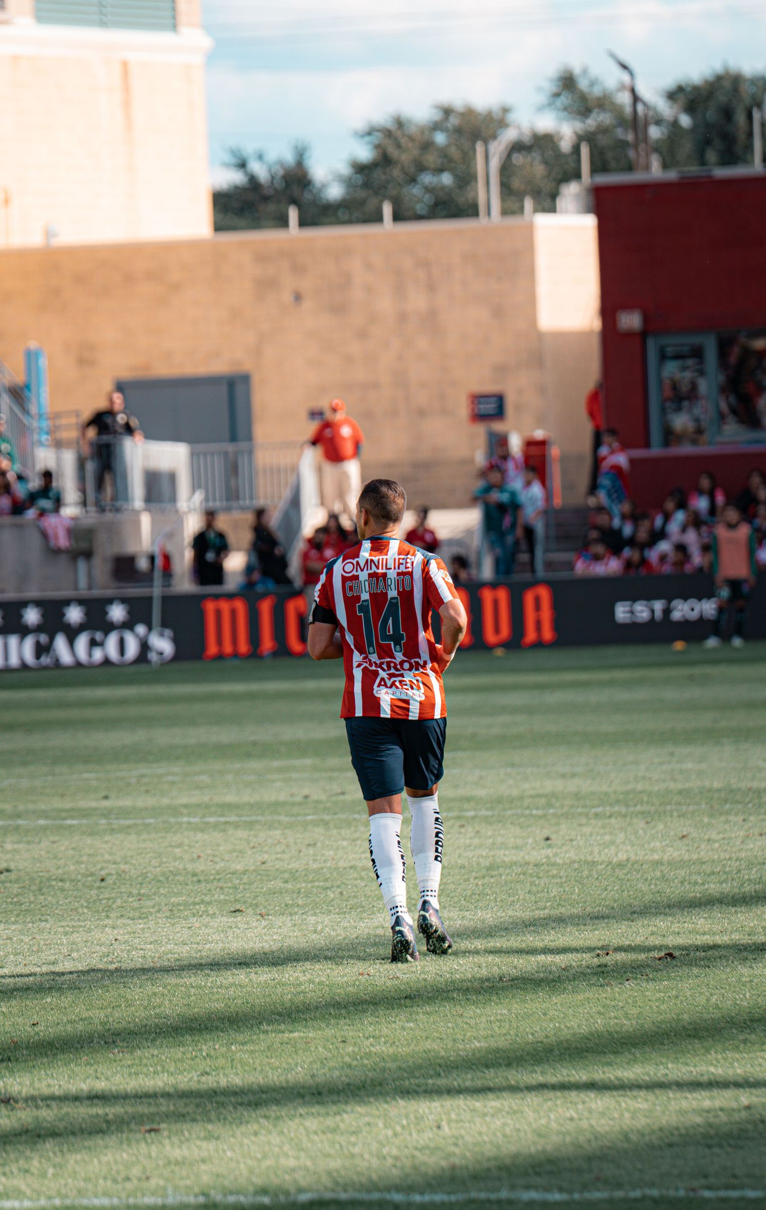 Players walking across sunny soccer field with stadium buildings visible in background.