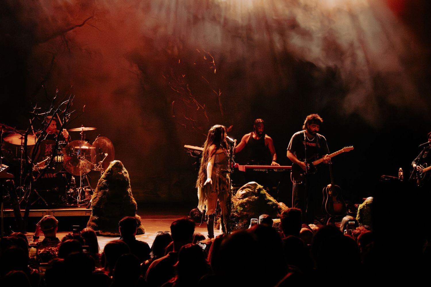 Silhouetted performers on a dark stage with dramatic red lighting create an atmospheric concert scene.