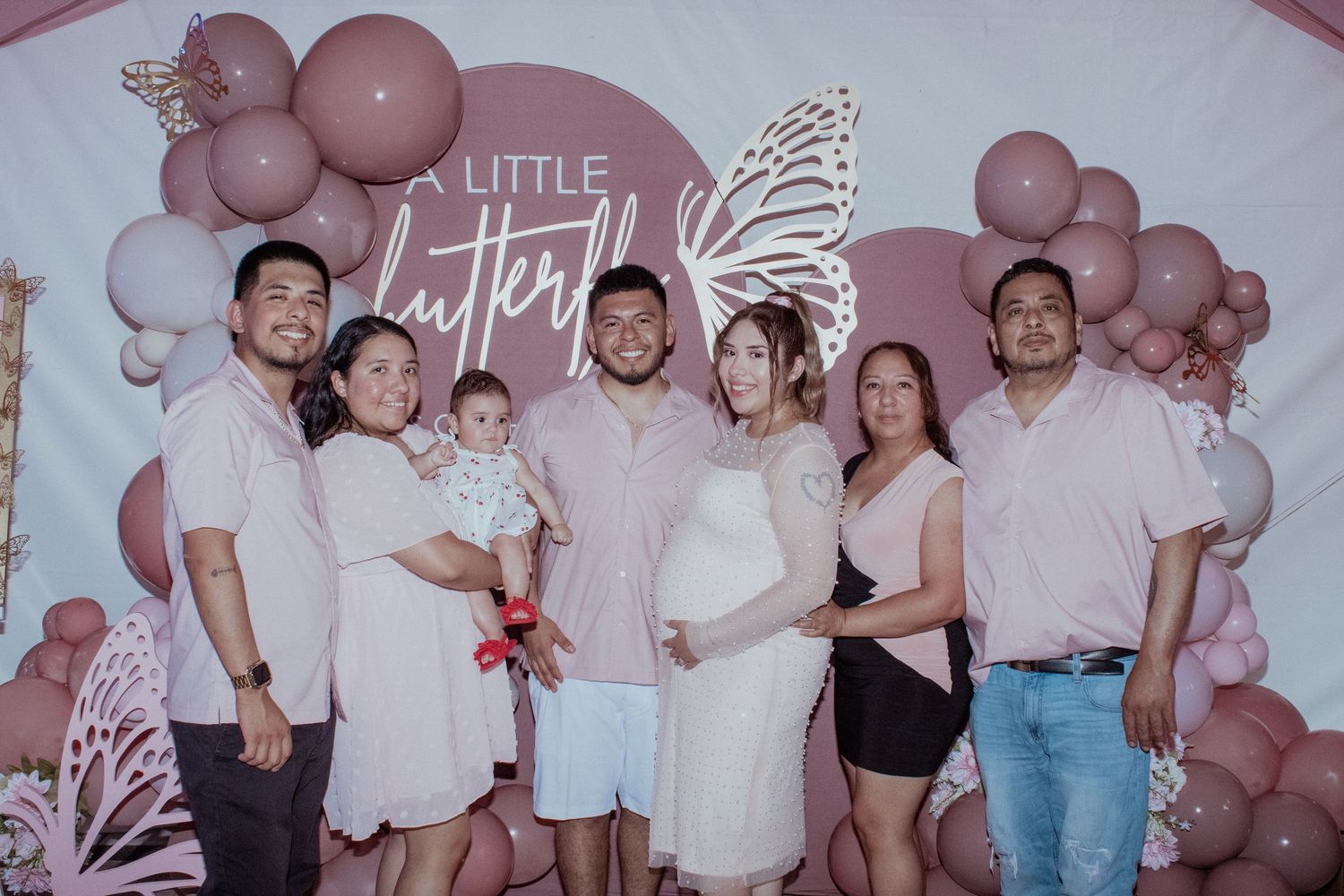 Group photo at a baby shower with pink balloon arch and white butterfly decorations.