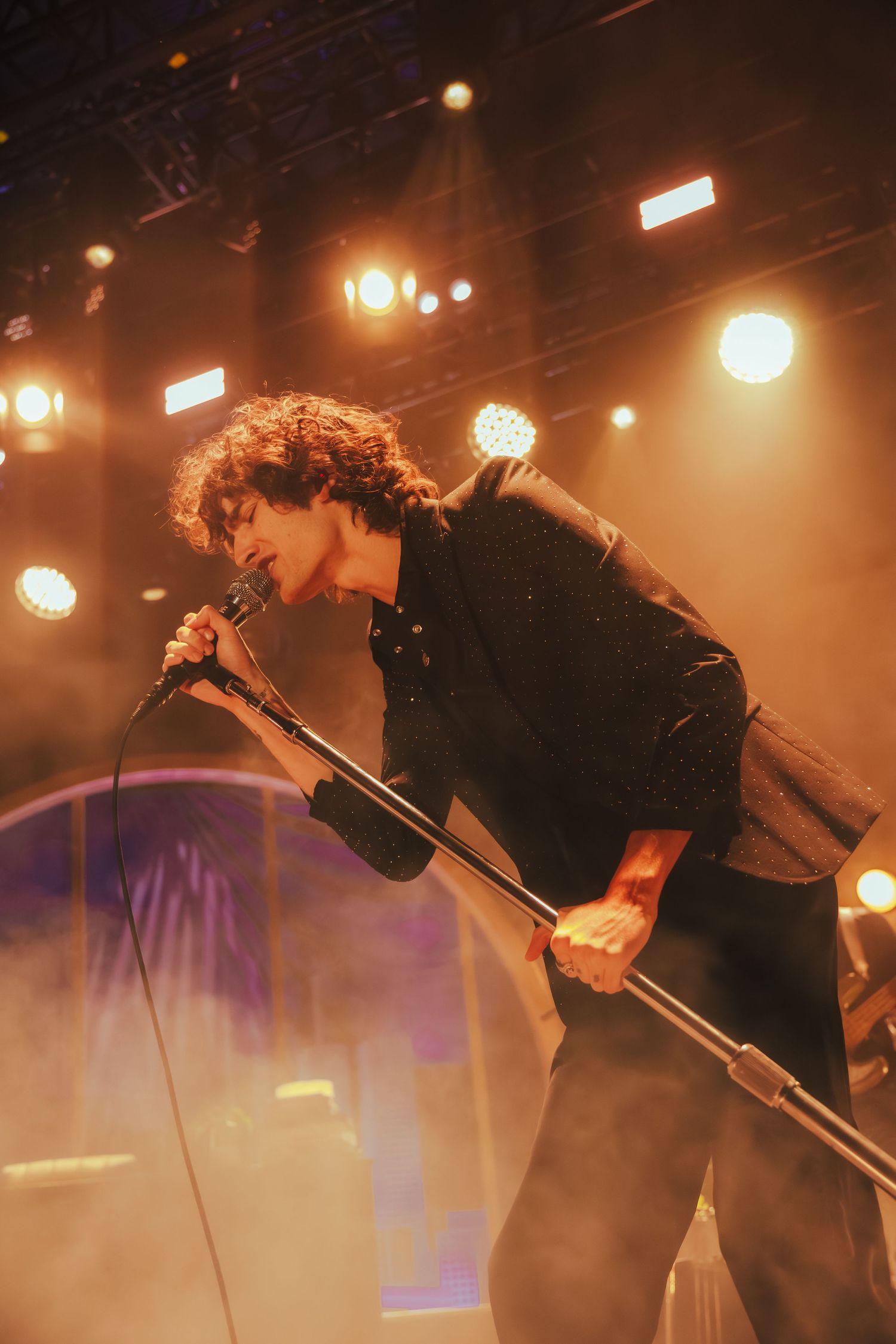Performer in dark clothing leans dramatically while singing on stage with warm lighting and atmospheric effects in background.