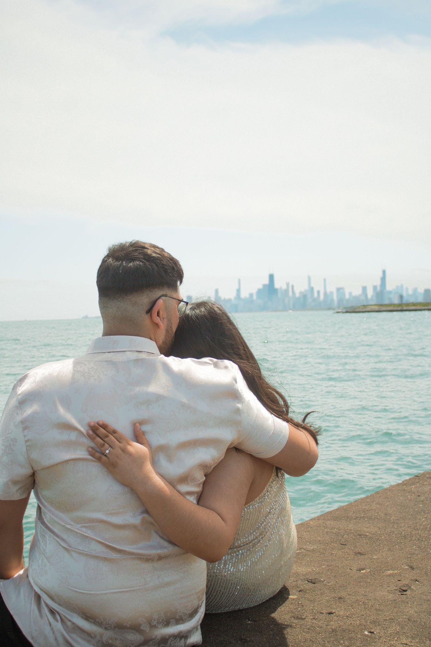 Two people embrace while sitting on a lakefront pier with a distant cityscape view.