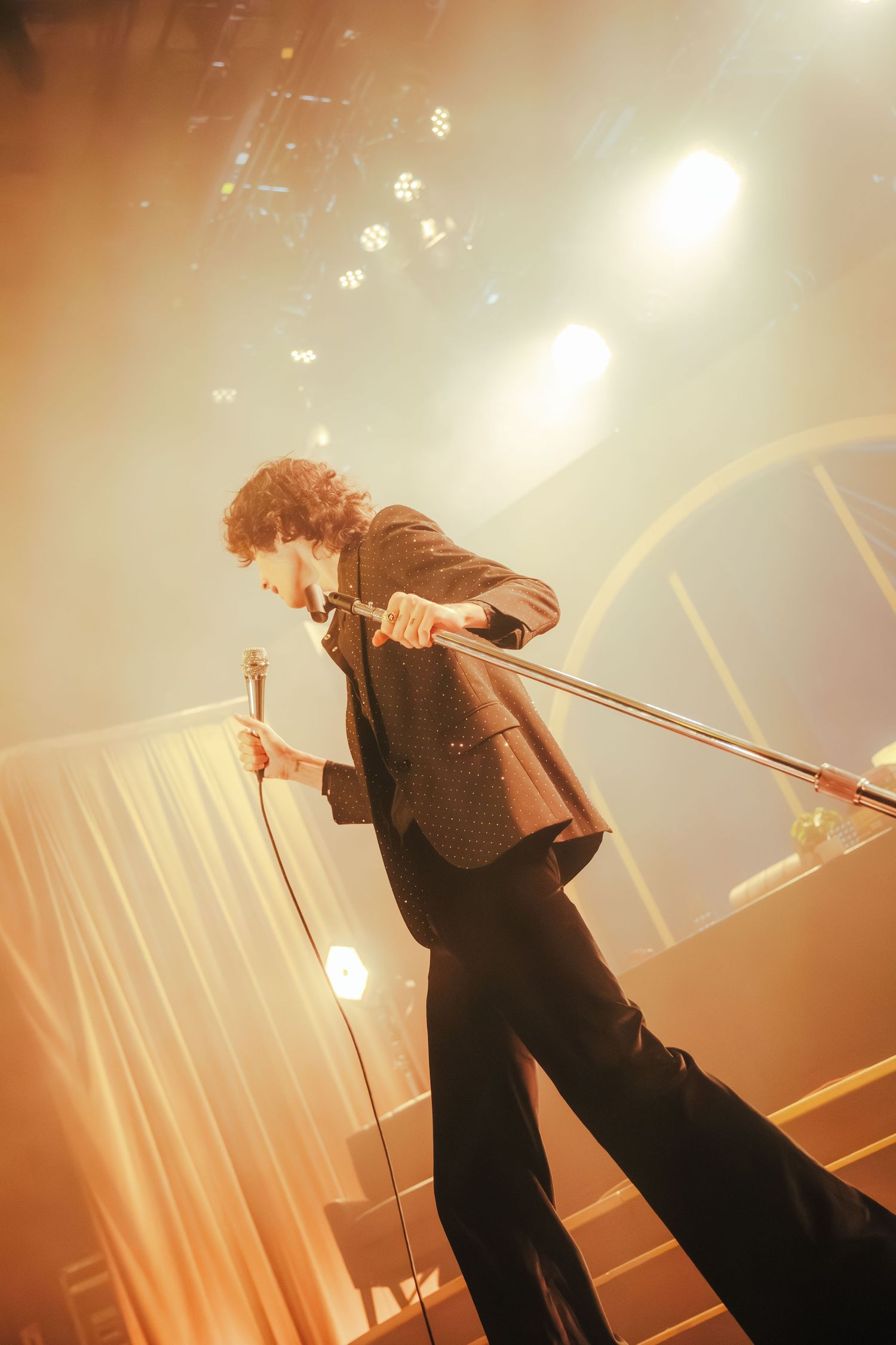 A performer in a brown suit jacket leans back dramatically on stage under warm spotlights during a concert performance.