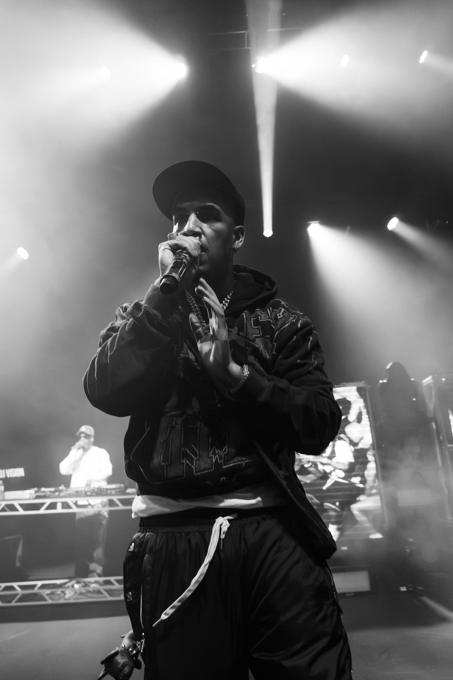 Hip hop artist performing on stage with stark white spotlights and DJ setup in background.