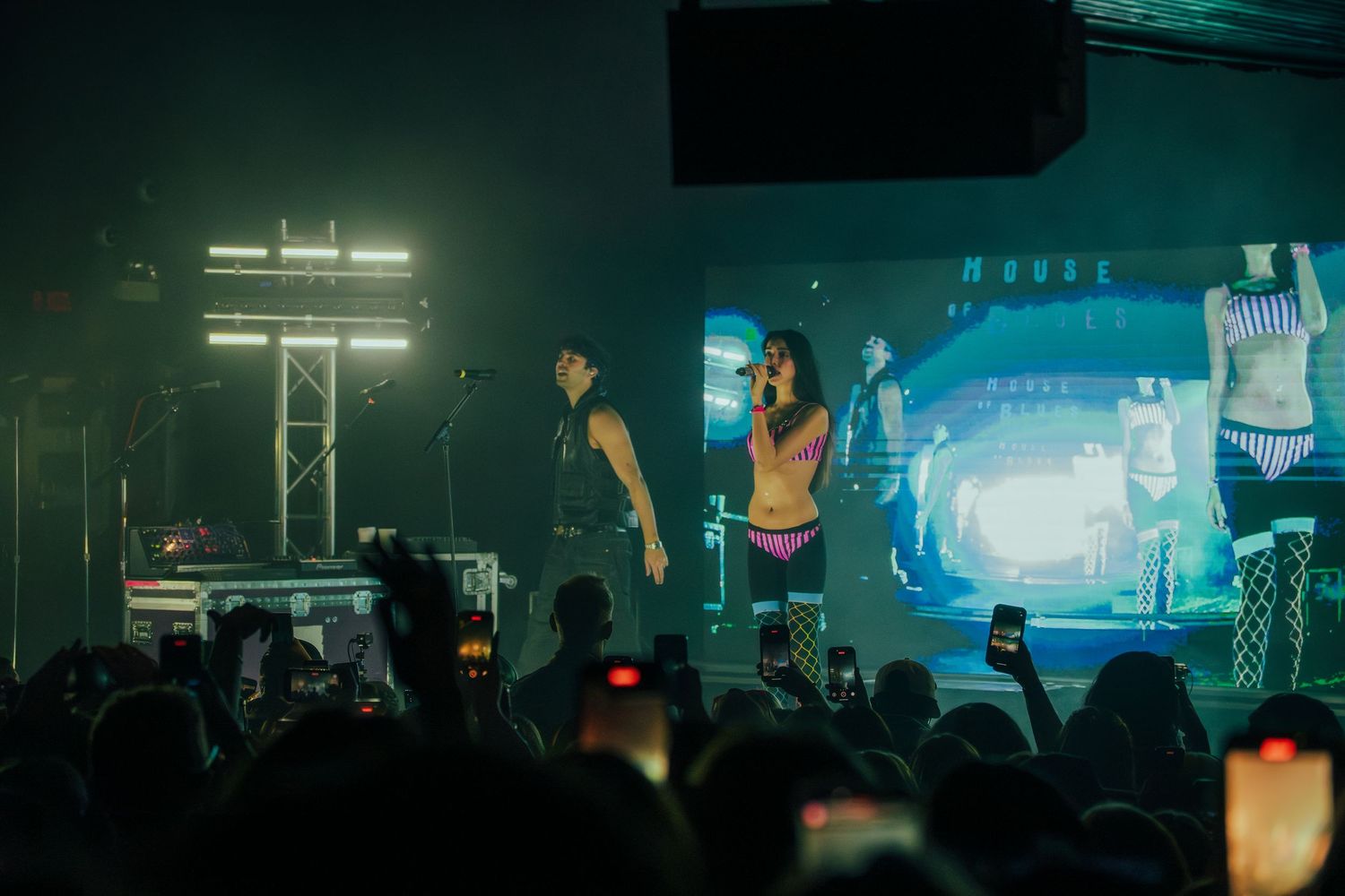 Concert performers on stage with dramatic blue and green lighting while crowd watches with phones raised in a dark venue.