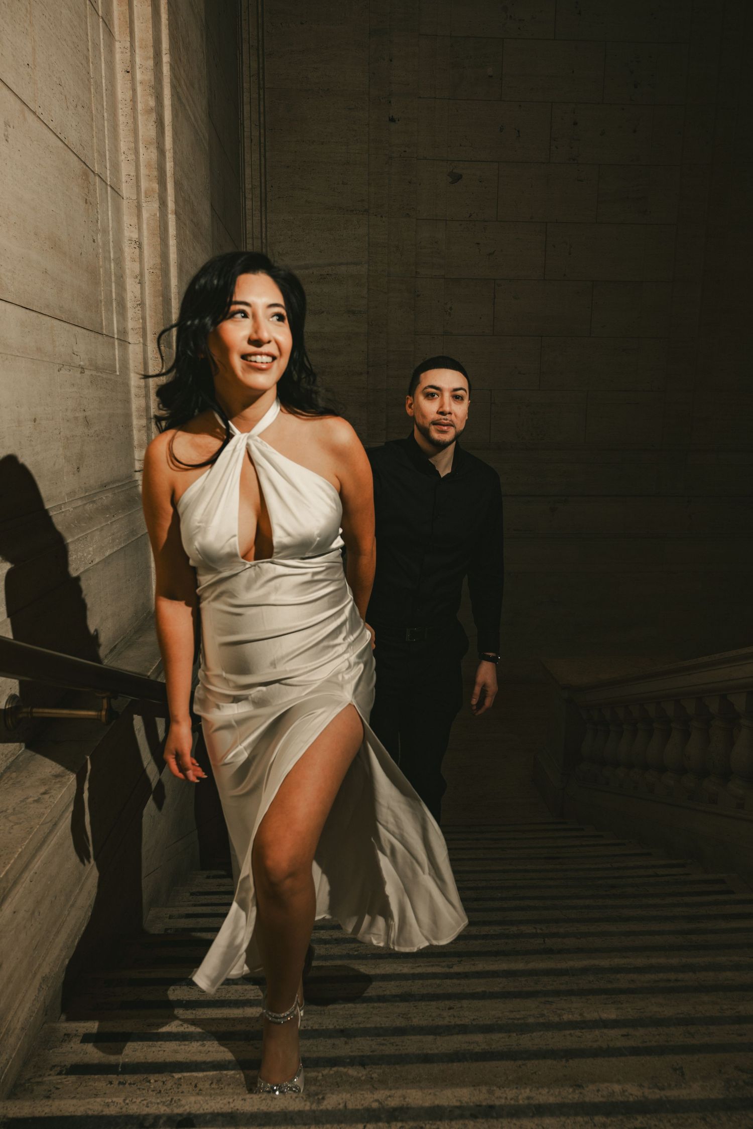 A person in a flowing white dress runs up stone stairs while someone in black follows behind.