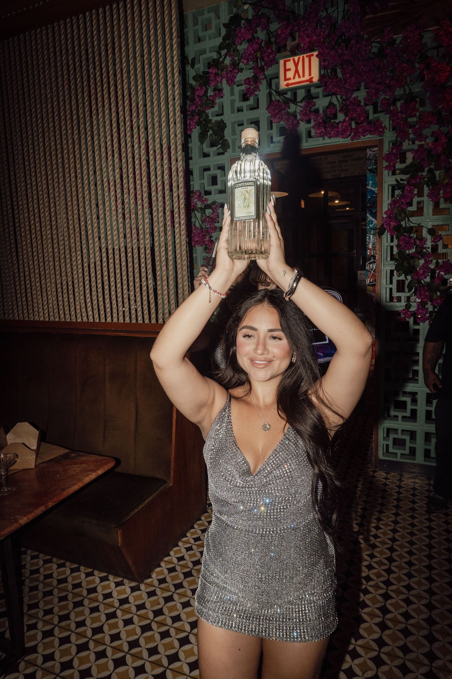 Person in shimmering silver top poses against floral wallpaper in a club setting.