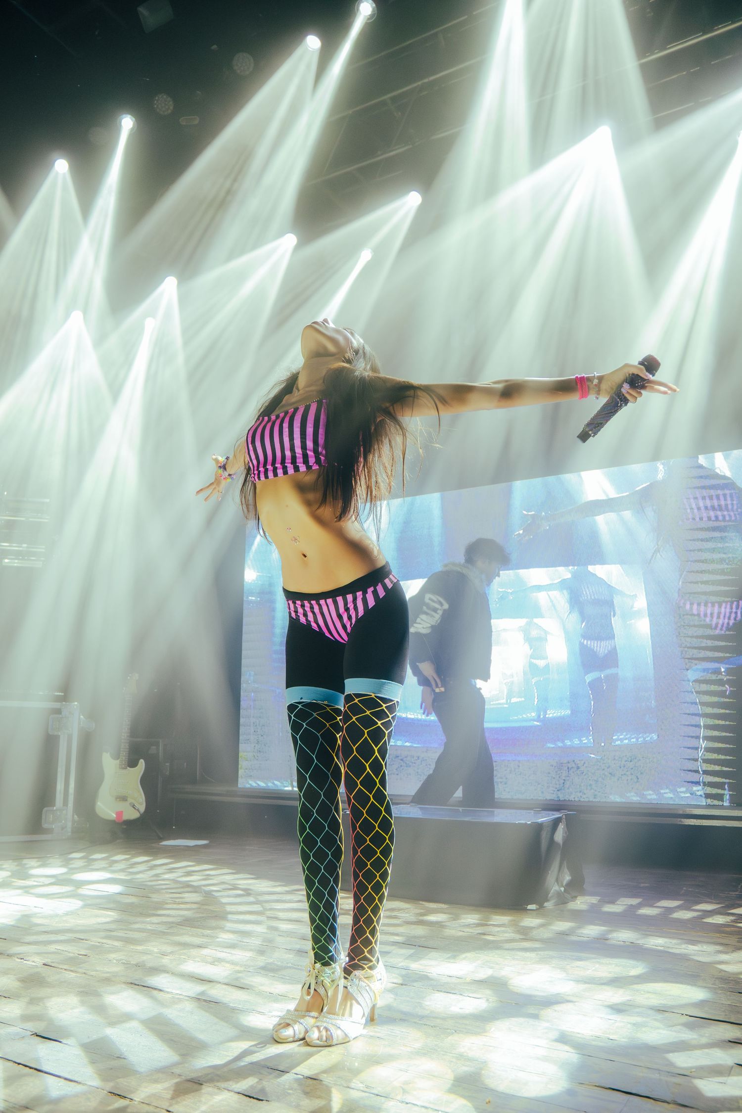 A performer in striped top and fishnet stockings dances on stage with dramatic white lighting beams in the background.