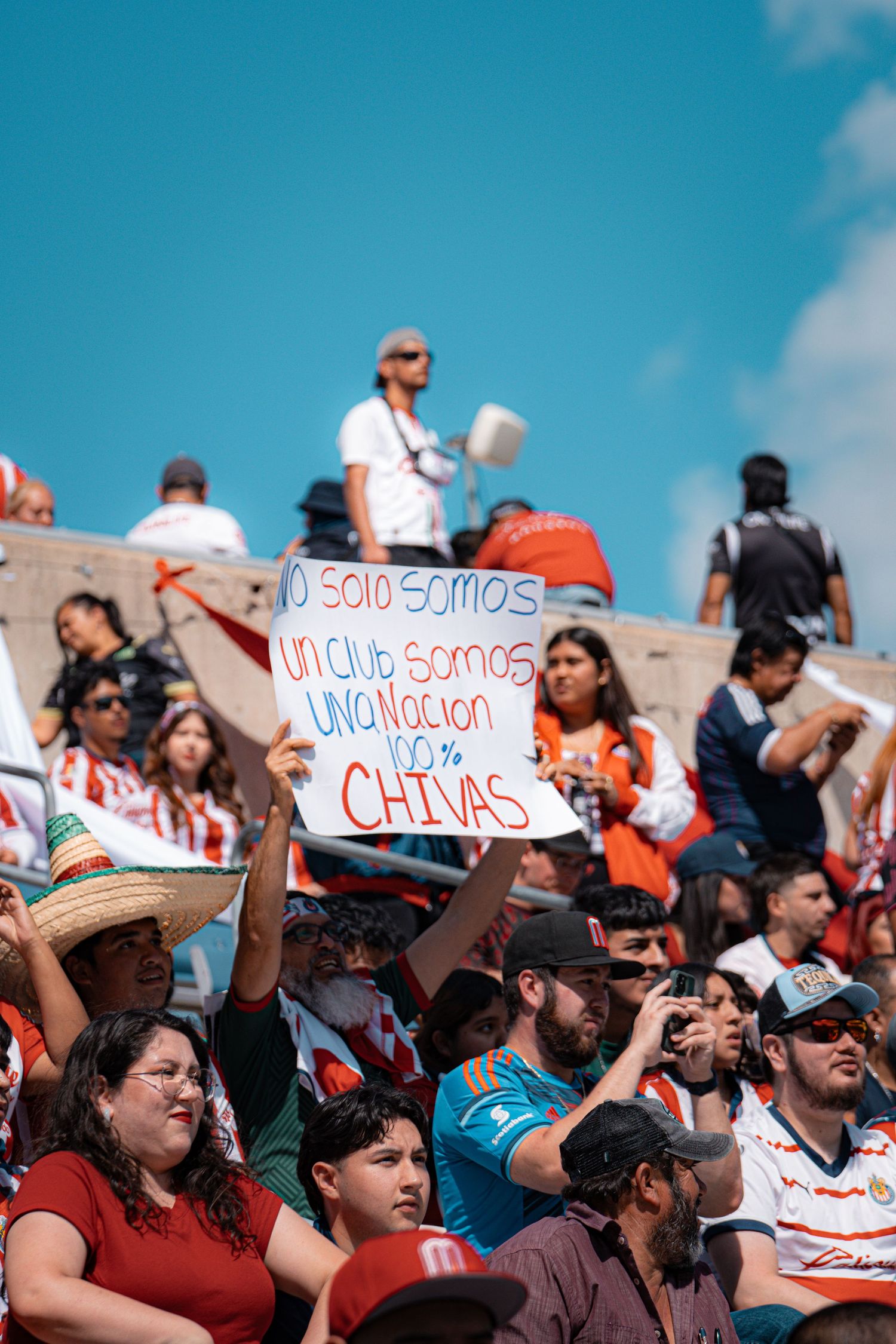 Passionate soccer fans hold up signs and banners in a crowded stadium under blue skies.