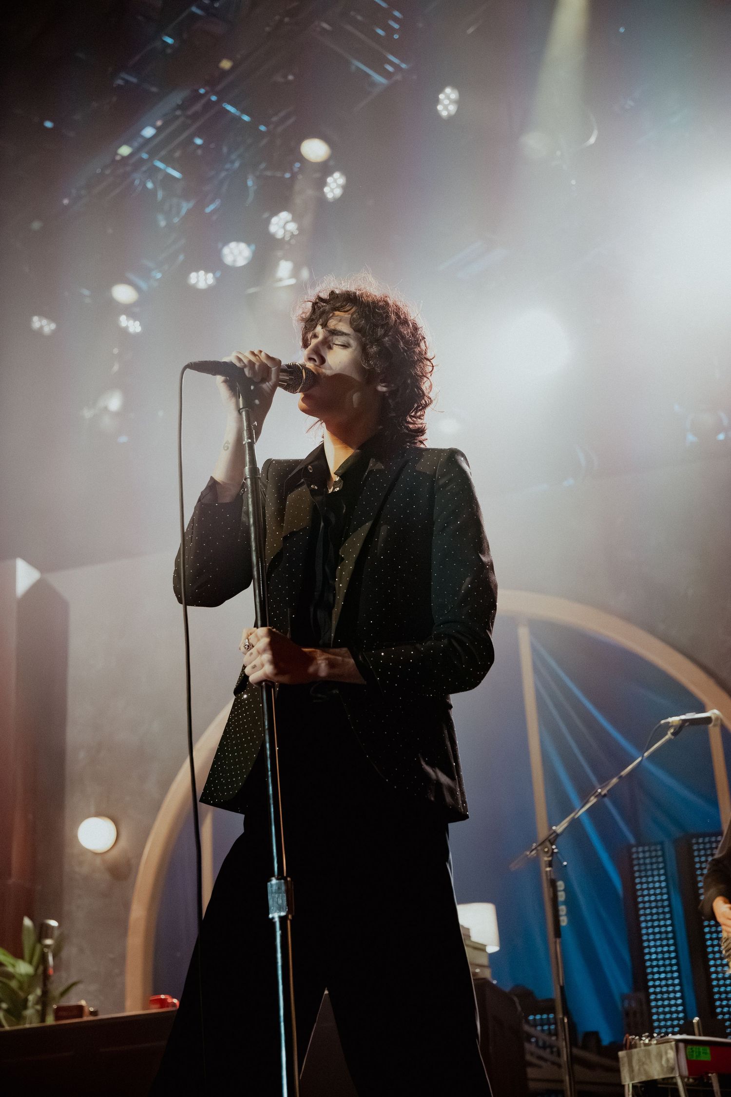 A singer performs on stage in dark lighting with a microphone stand and atmospheric fog effects creating a moody ambiance.