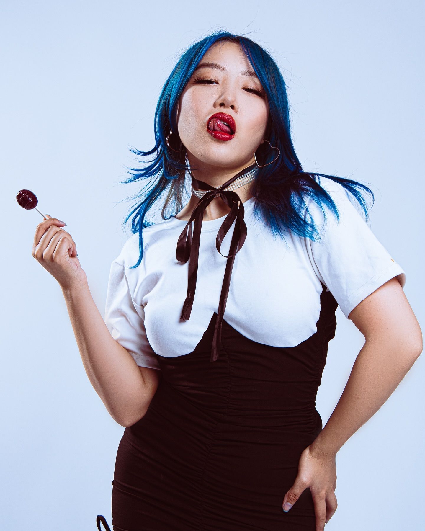 Fashion portrait of a model with blue hair holding a lollipop in a black and white outfit.