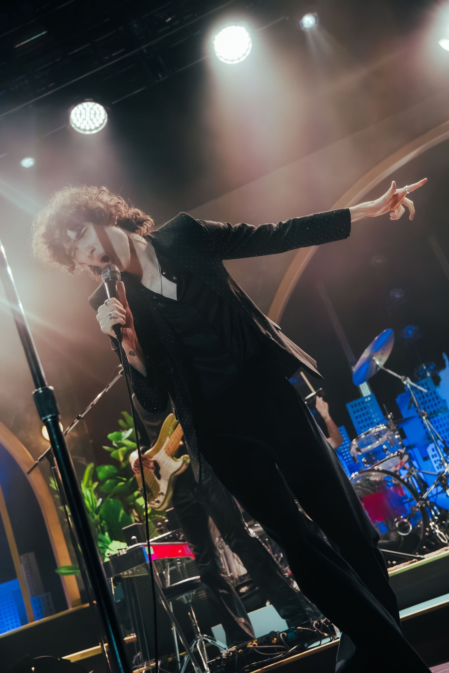 A performer in black clothing strikes dramatic poses on stage with blue lighting and musical equipment in the background.
