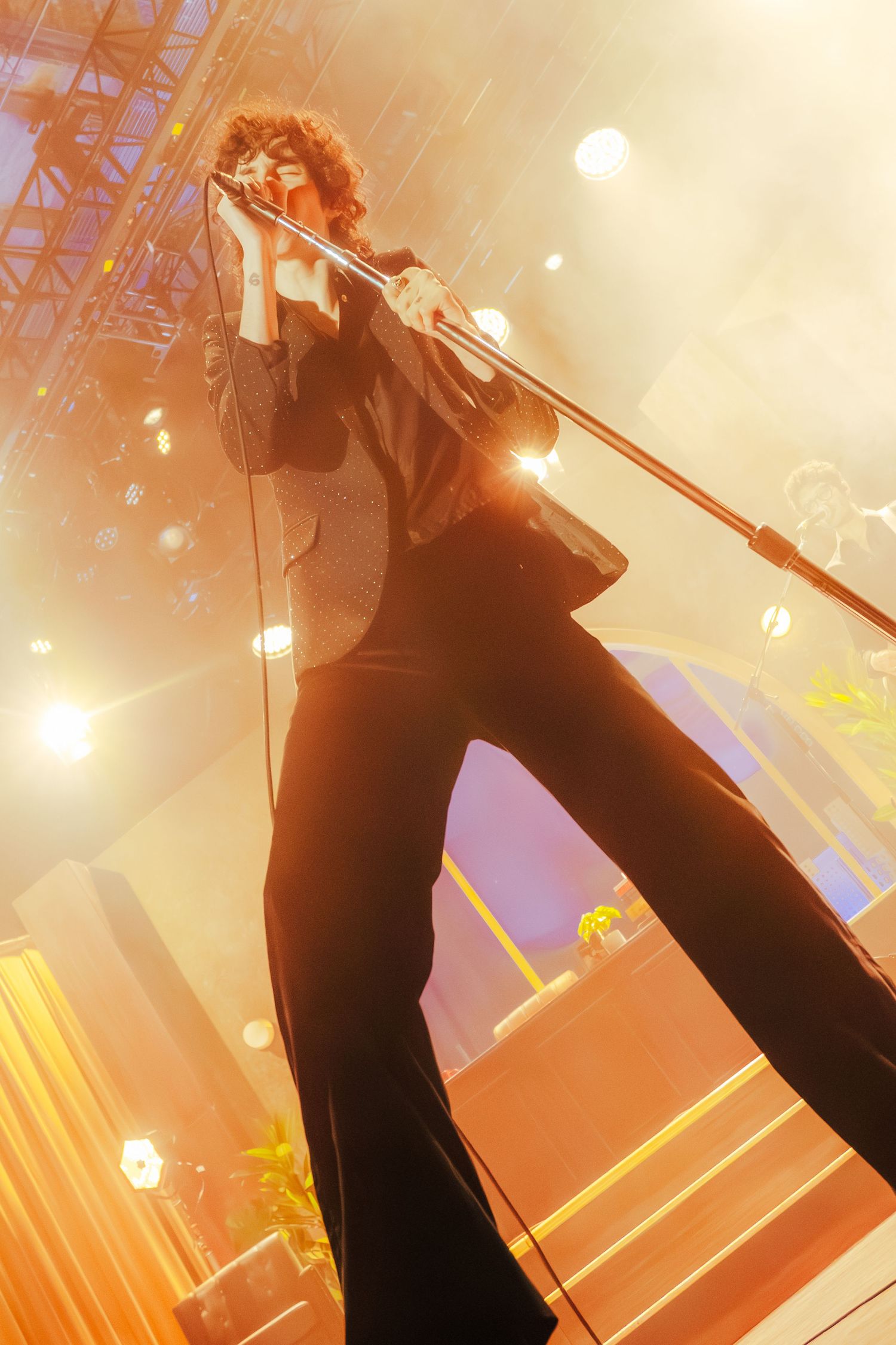 Performer in black suit silhouetted against orange stage lights leans back while singing into microphone stand during concert.