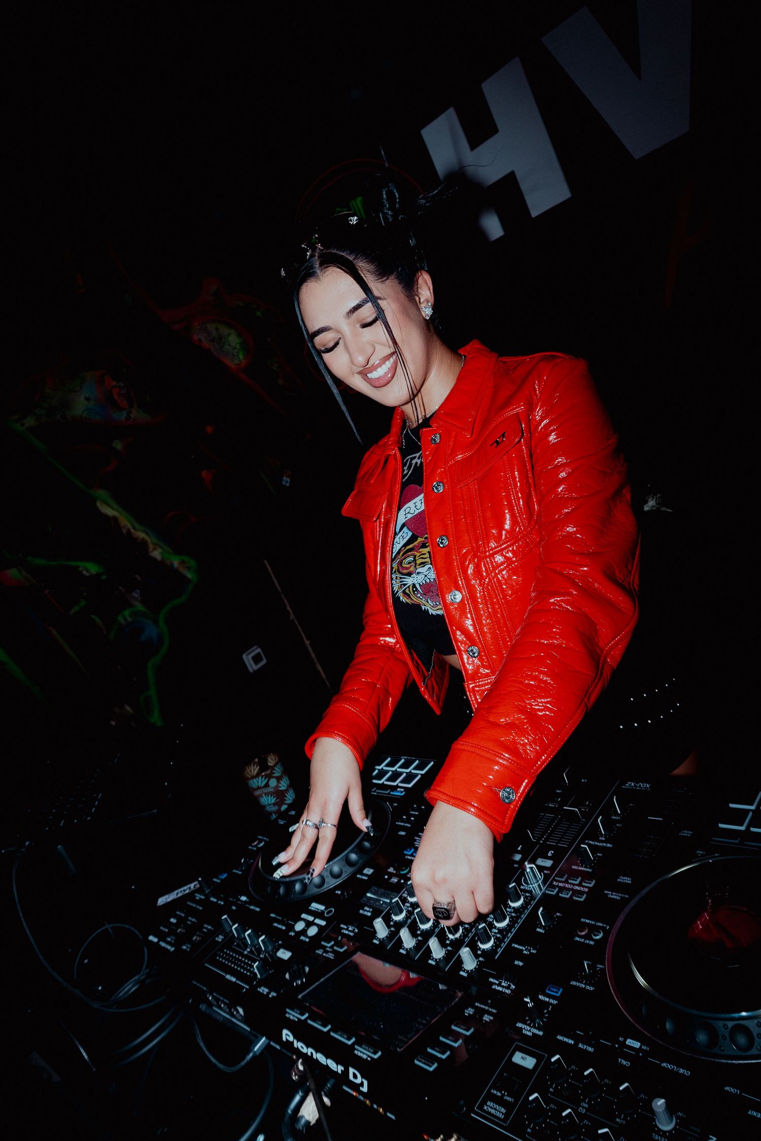 DJ in a bright red jacket mixing music on turntables in dark club setting.