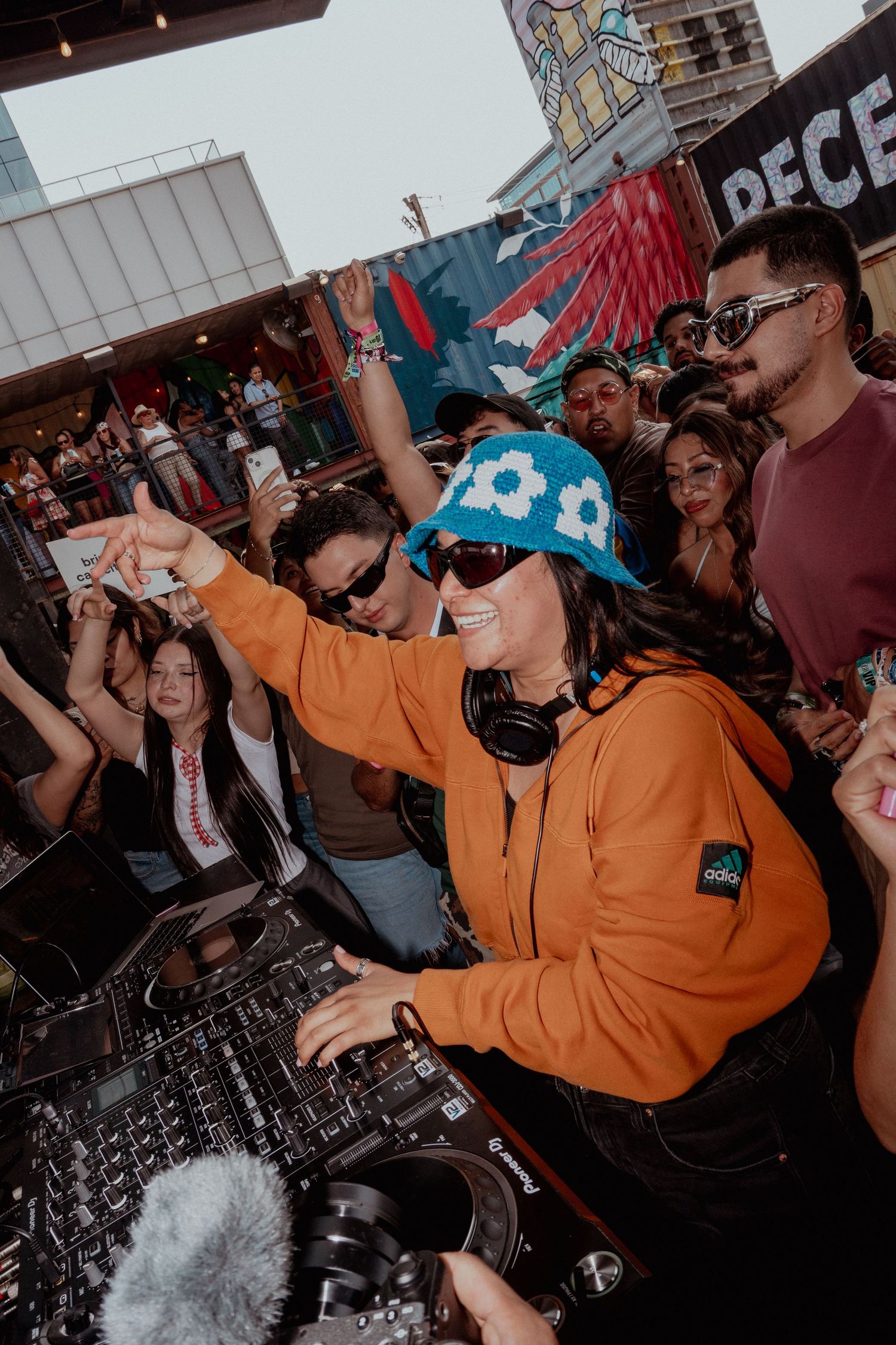 DJ in orange hoodie performing energetically at a crowded event with mixing equipment.