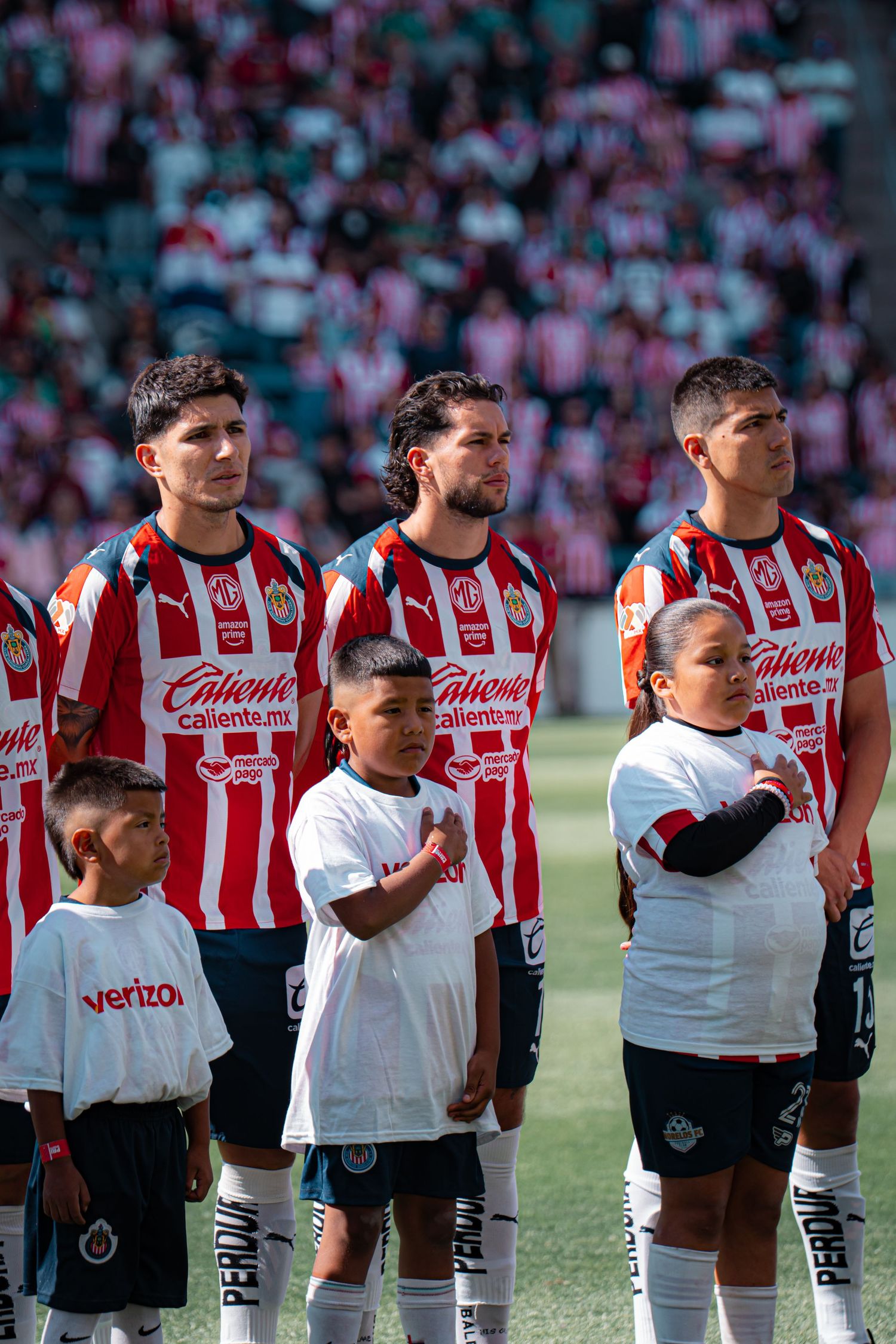 Soccer players and young escorts stand in formation before a match wearing red and white striped jerseys.