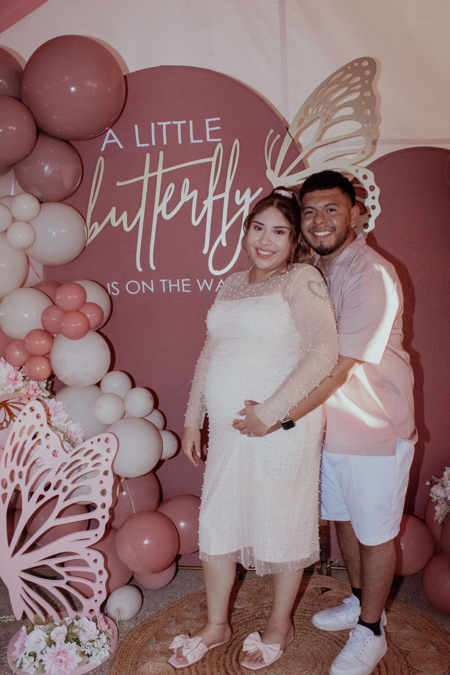 Baby shower celebration with pink balloon arch and butterfly decorations against a mauve backdrop.