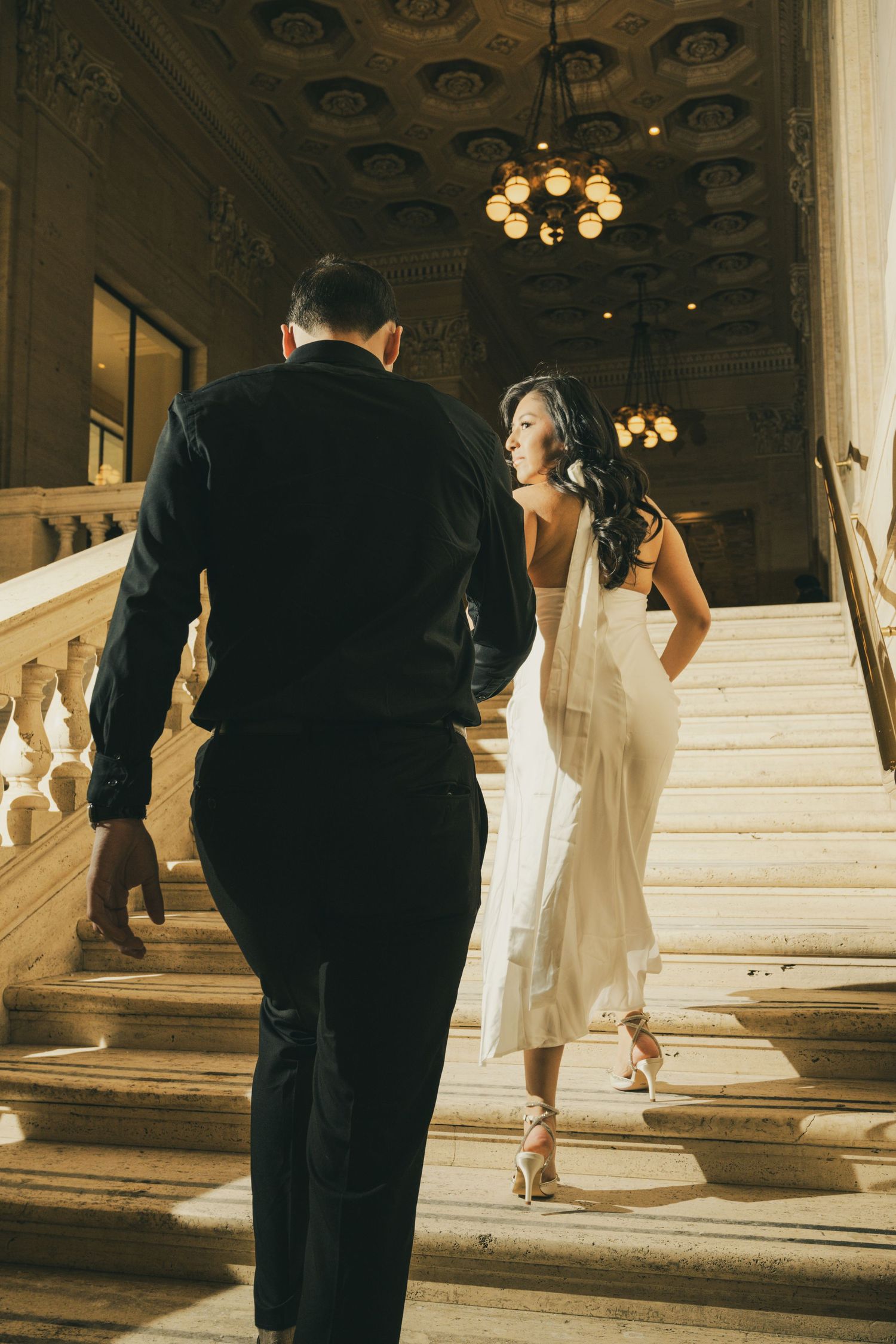 A couple in formal attire walks up an elegant marble staircase in a grand venue with ornate lighting.