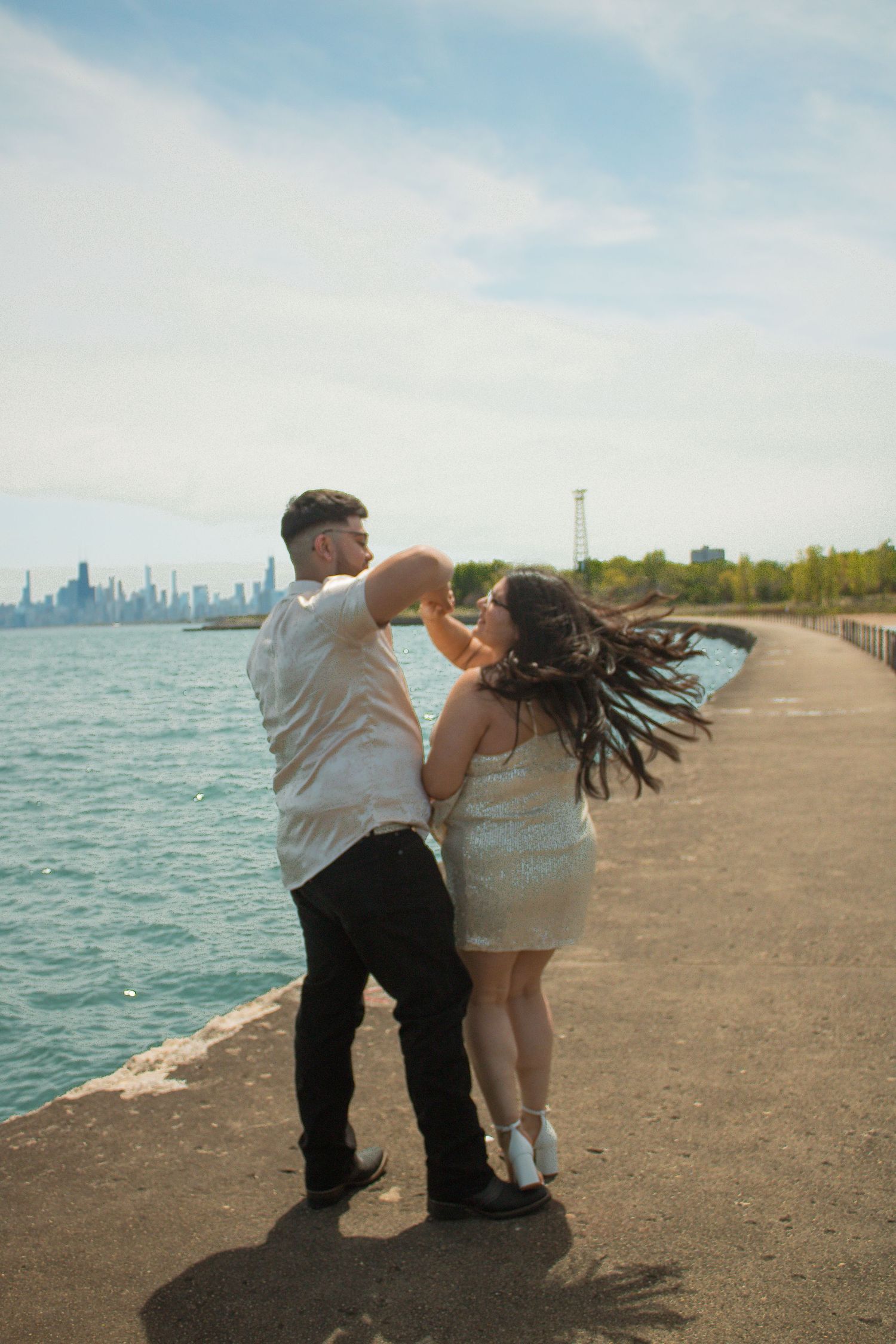 Couple poses playfully on lakefront path with city skyline visible in background.