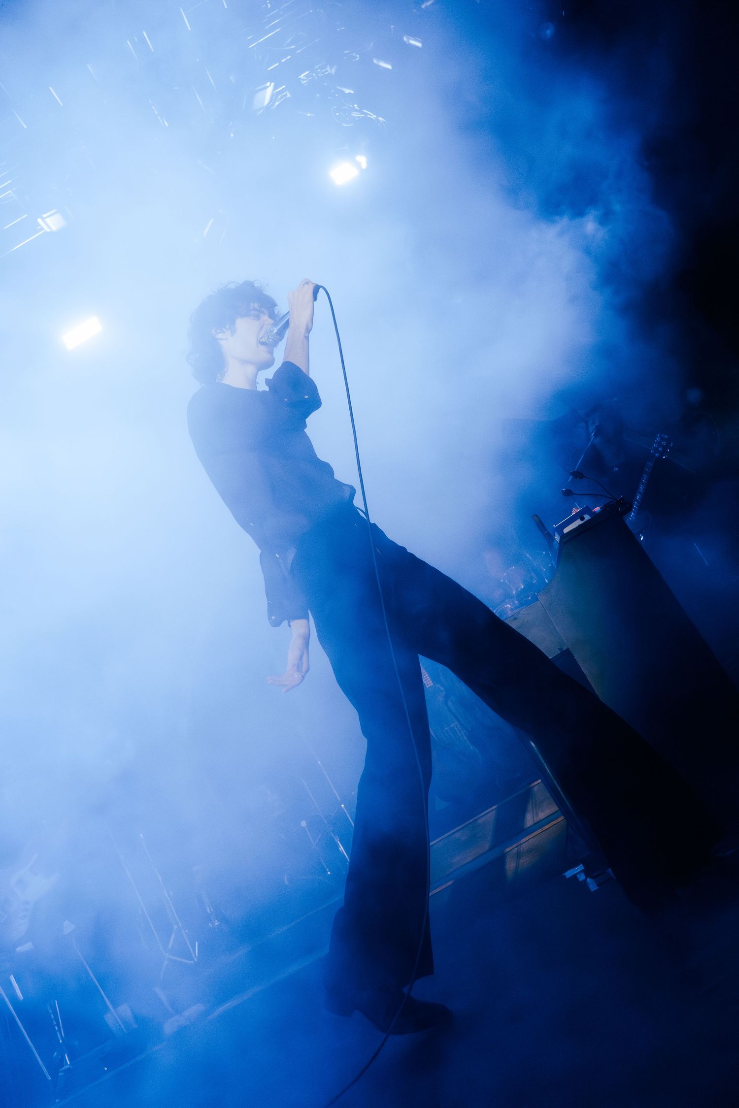 Silhouetted figures perform on a dark stage with dramatic blue smoke and lighting effects creating an atmospheric scene.