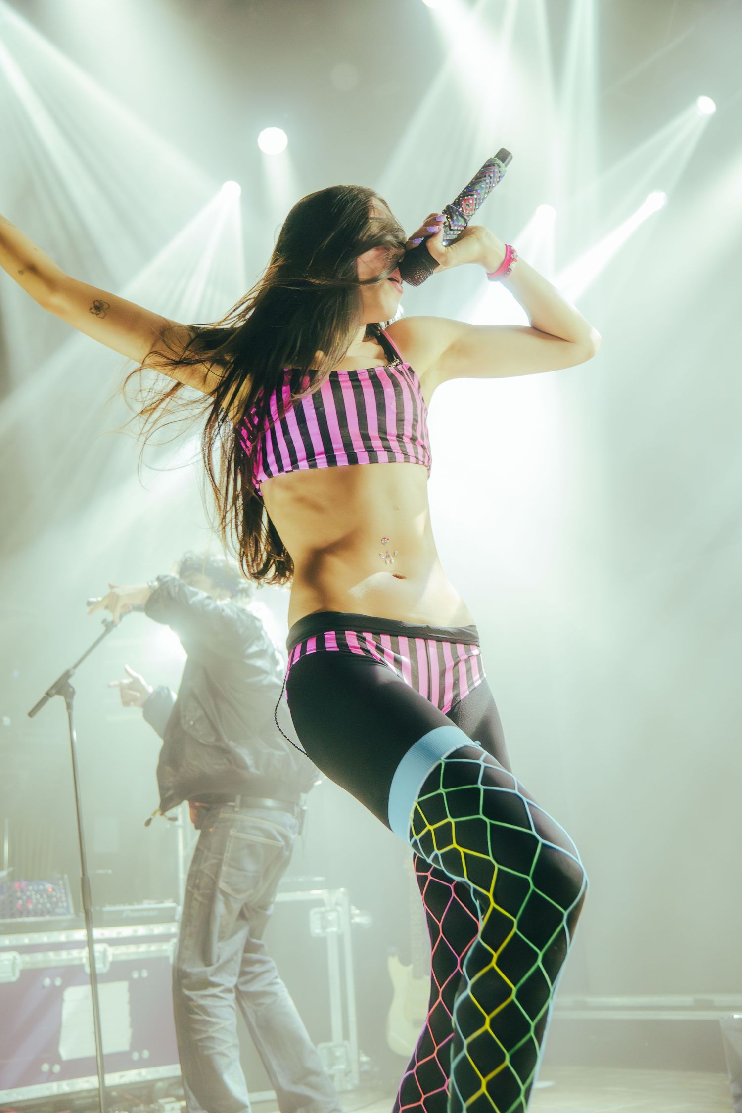 Performer in striped top and fishnet stockings dances energetically on stage with dramatic concert lighting in background.