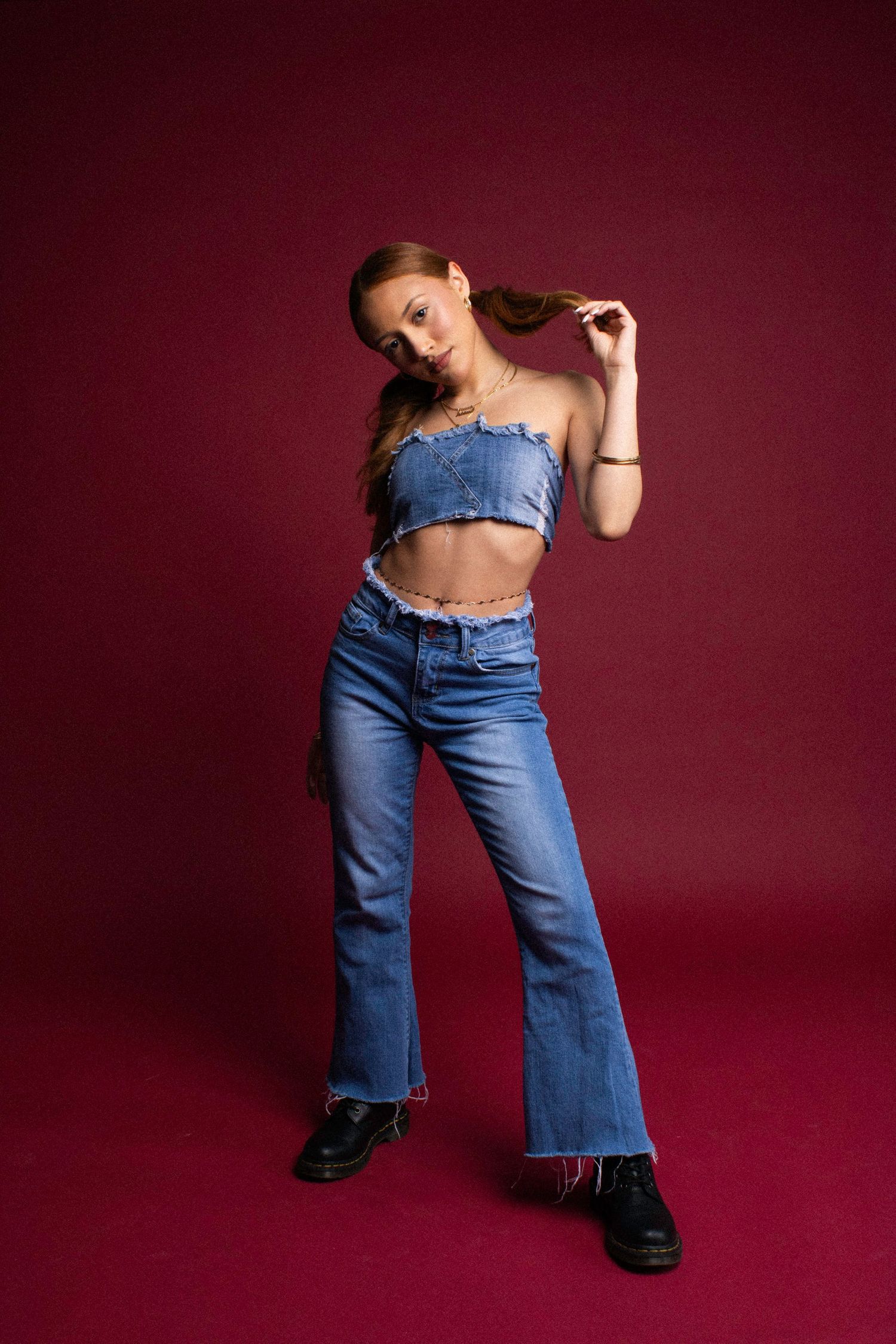 Model poses in retro bell bottom jeans and black boots against a burgundy backdrop in fashion photoshoot series.