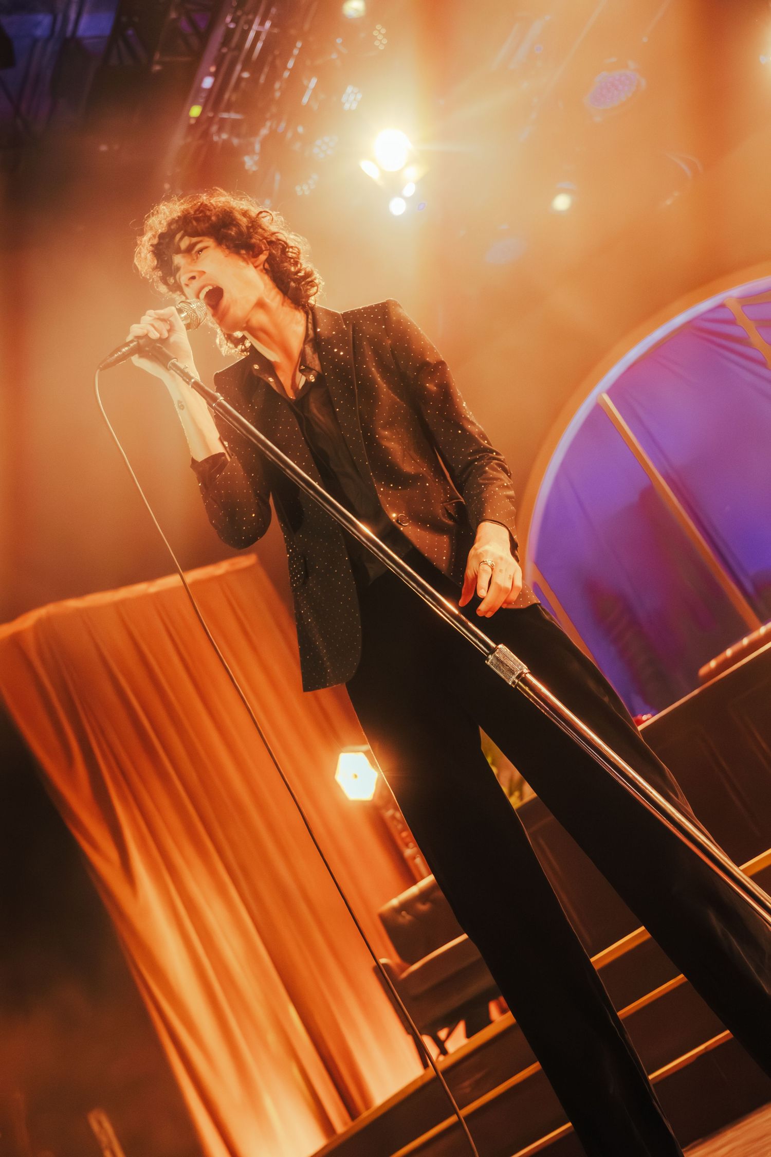 Performer in black attire sings passionately on stage with dramatic lighting and fog effects during live concert performance.