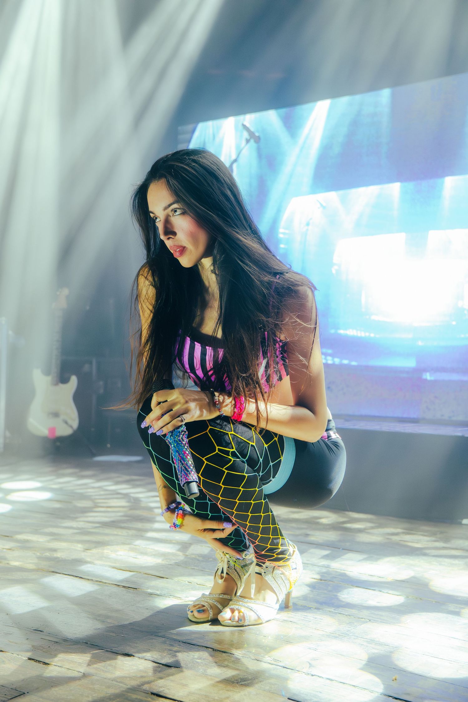 A person in colorful patterned leggings and platform shoes crouches on a reflective floor with dramatic lighting effects.