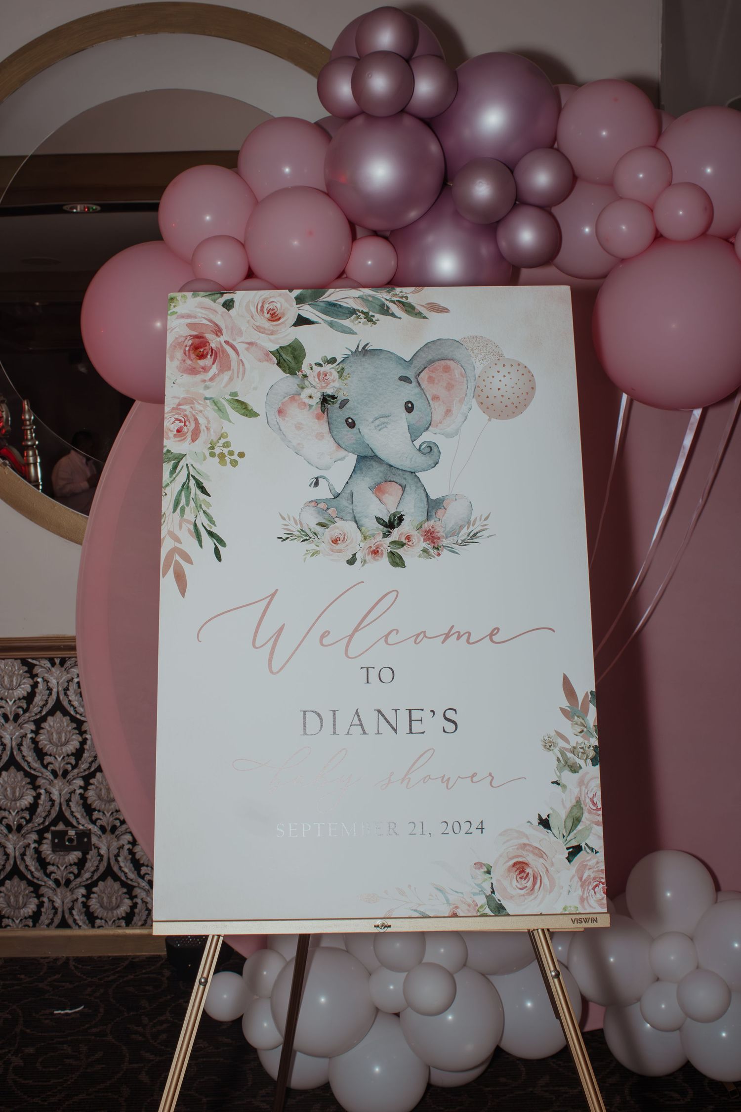 Welcome sign with floral design and elephant motif surrounded by pink and purple balloon arch.
