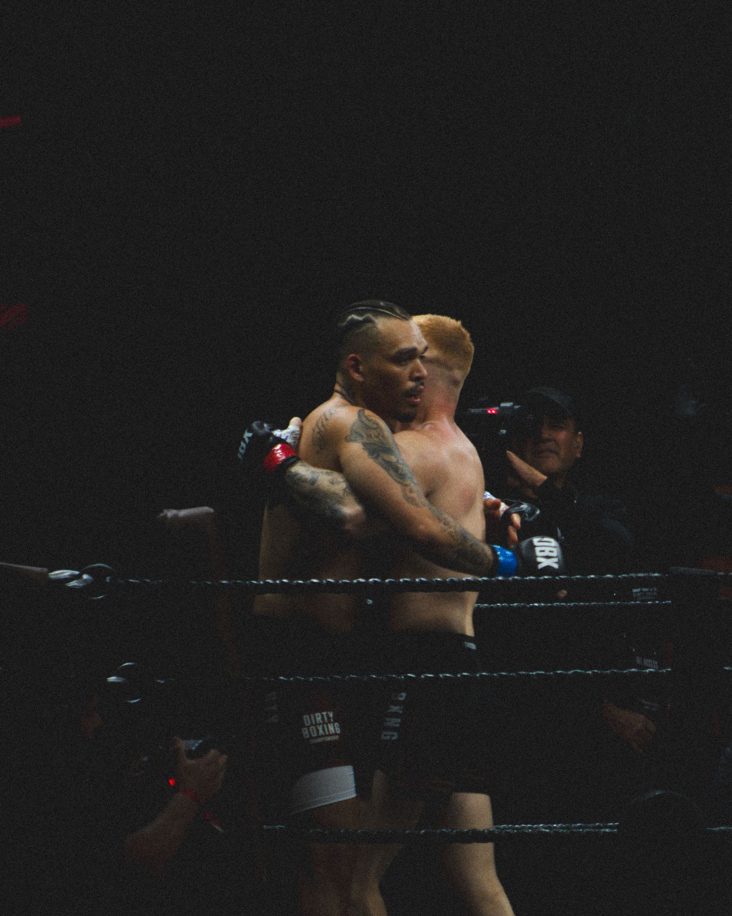Two fighters clinch against the ropes during an intense boxing match under dramatic lighting.