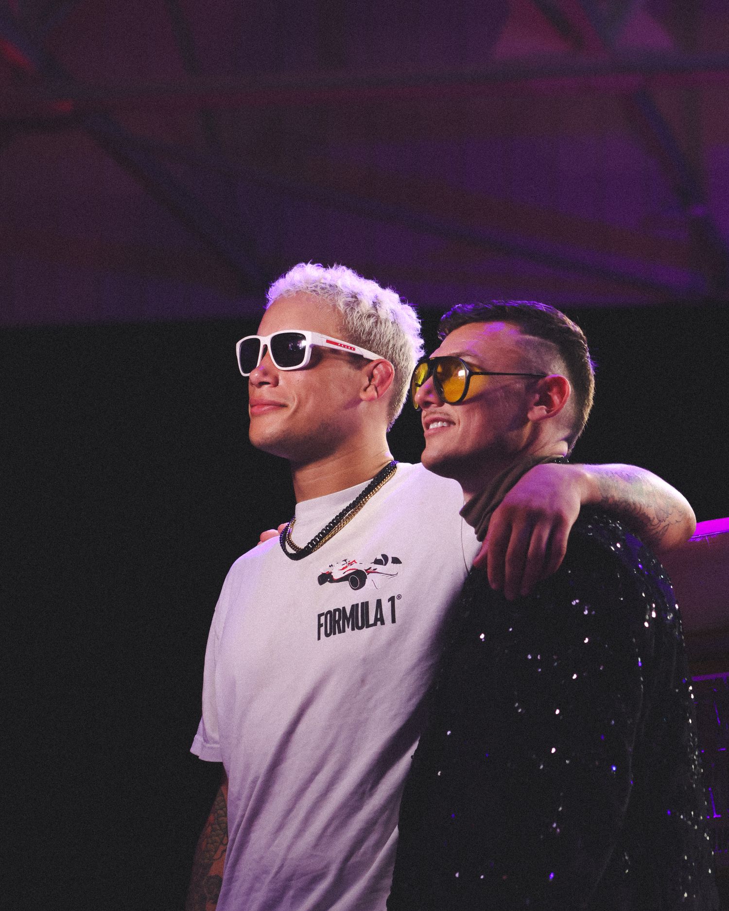 Two performers in sunglasses smile together under purple stage lighting.