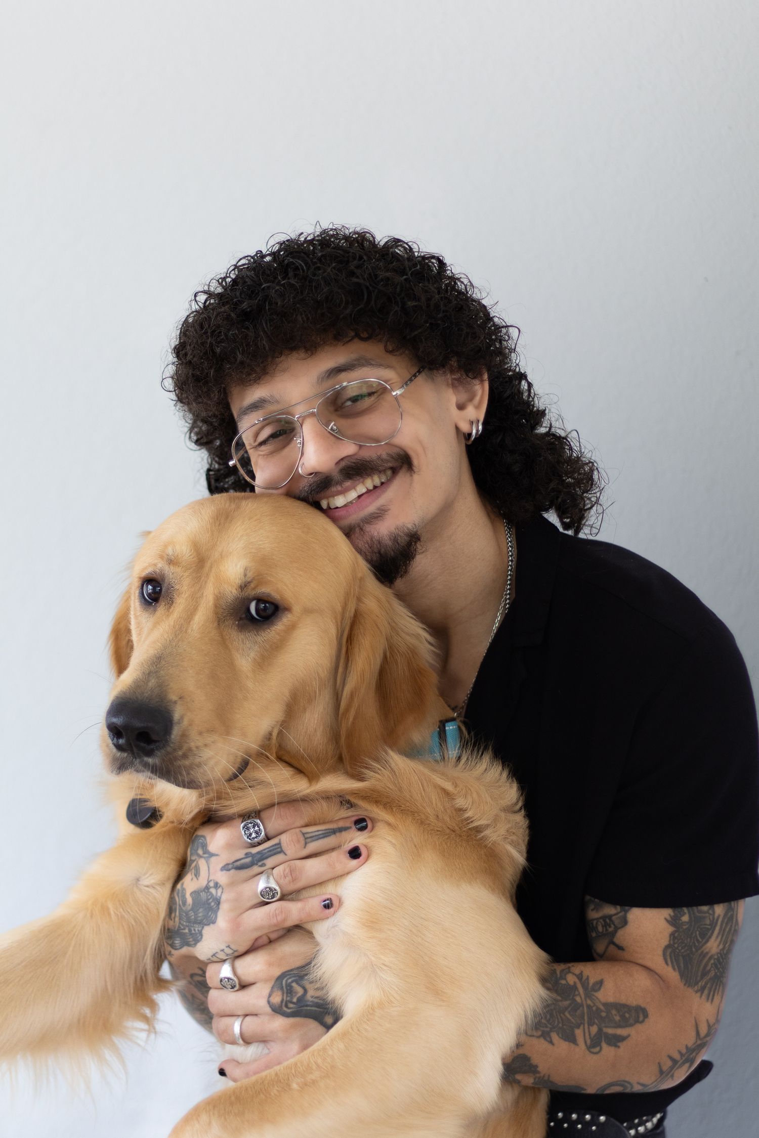 A person with curly hair and tattoos hugs and plays with a golden retriever dog against a light background.