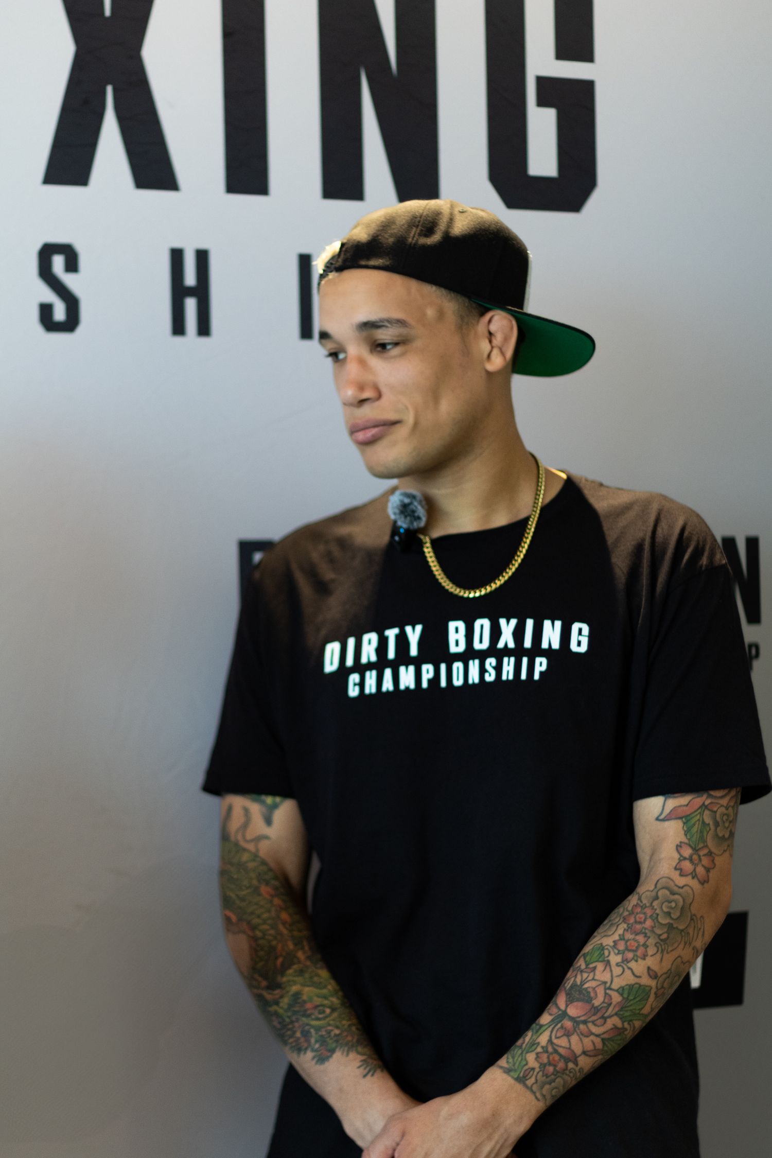 Person in black championship shirt with tattoos speaking during boxing event.