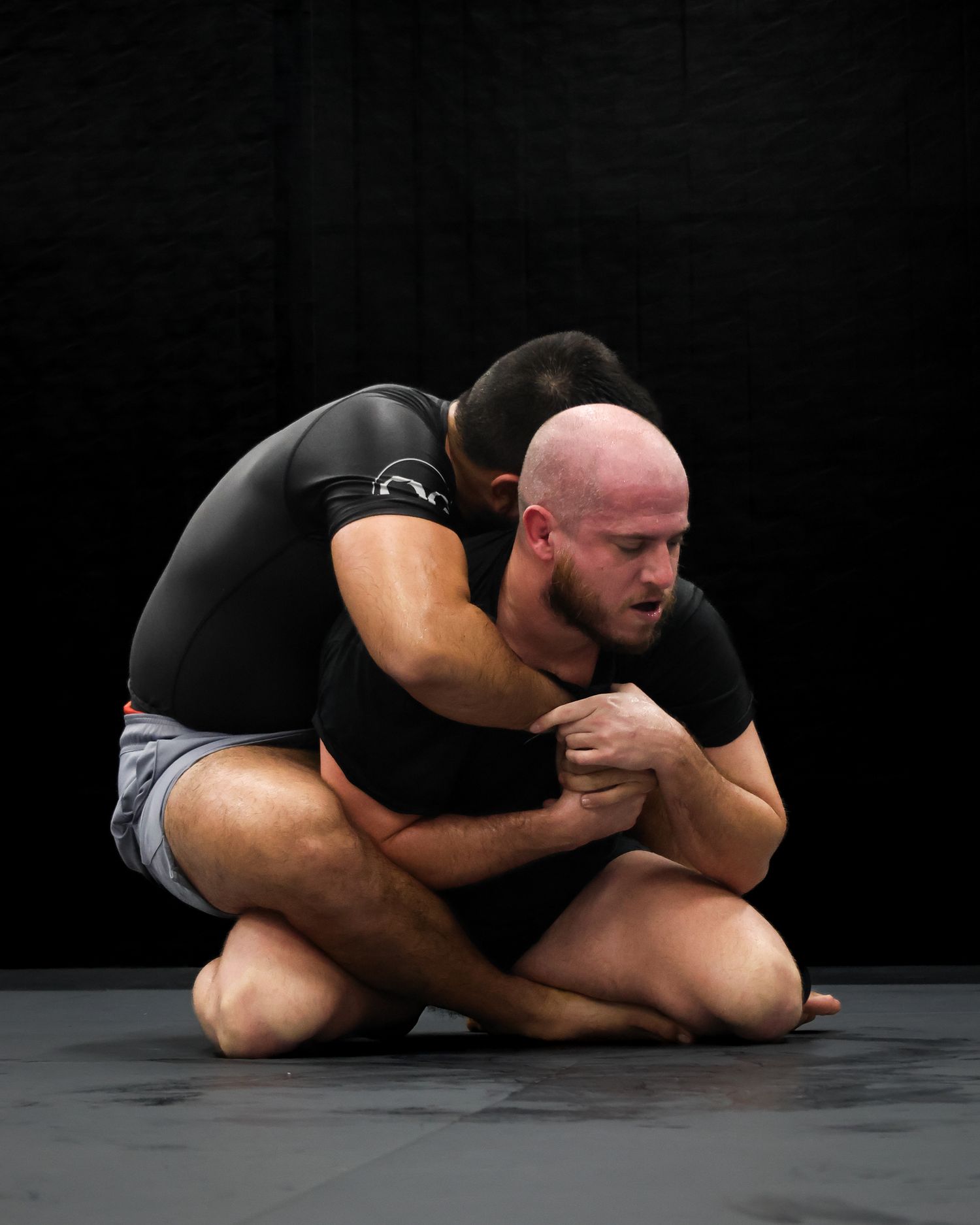 Two grapplers demonstrate wrestling hold techniques on dark mat.