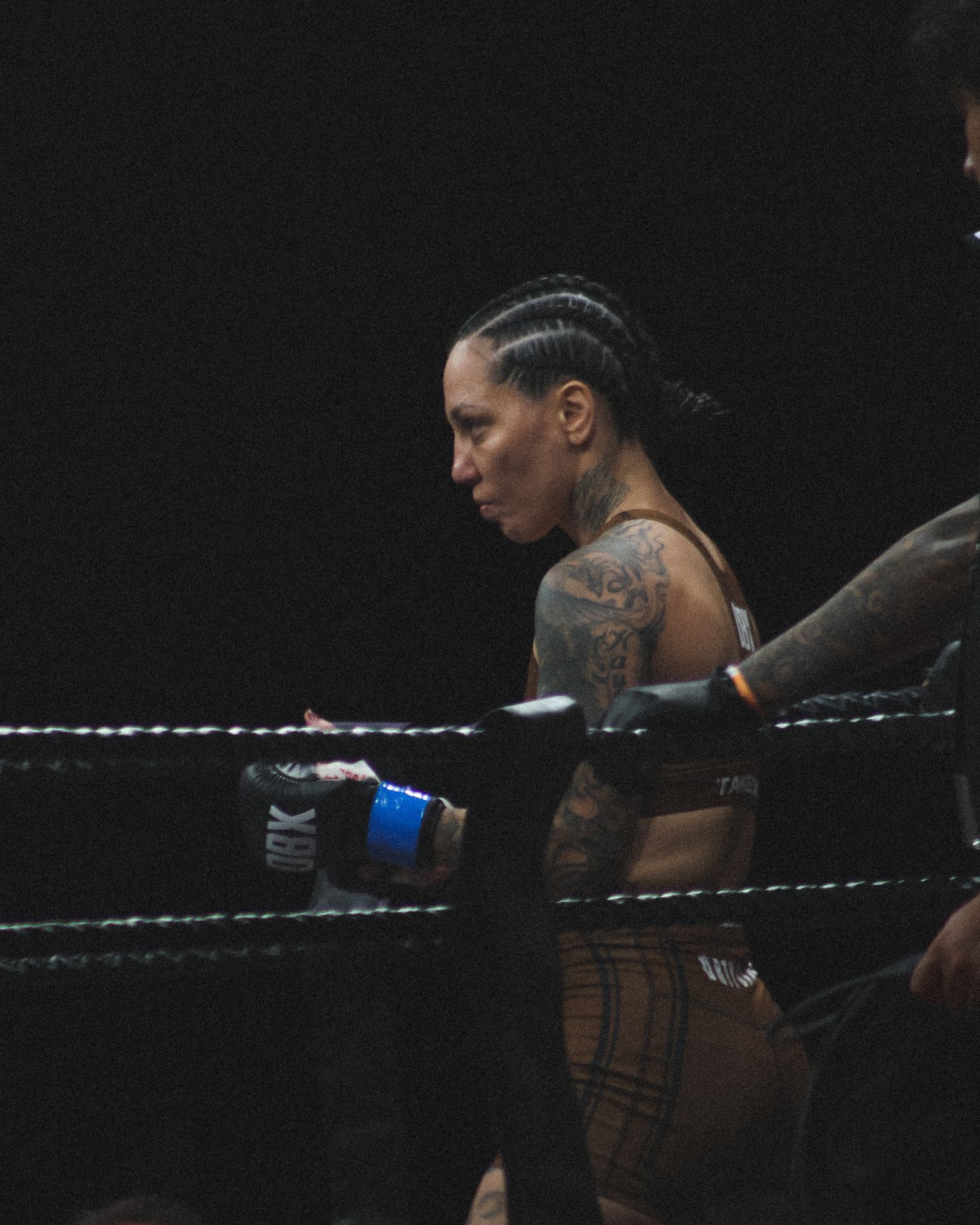 Female wrestler in black athletic wear leans against ring ropes during professional match.