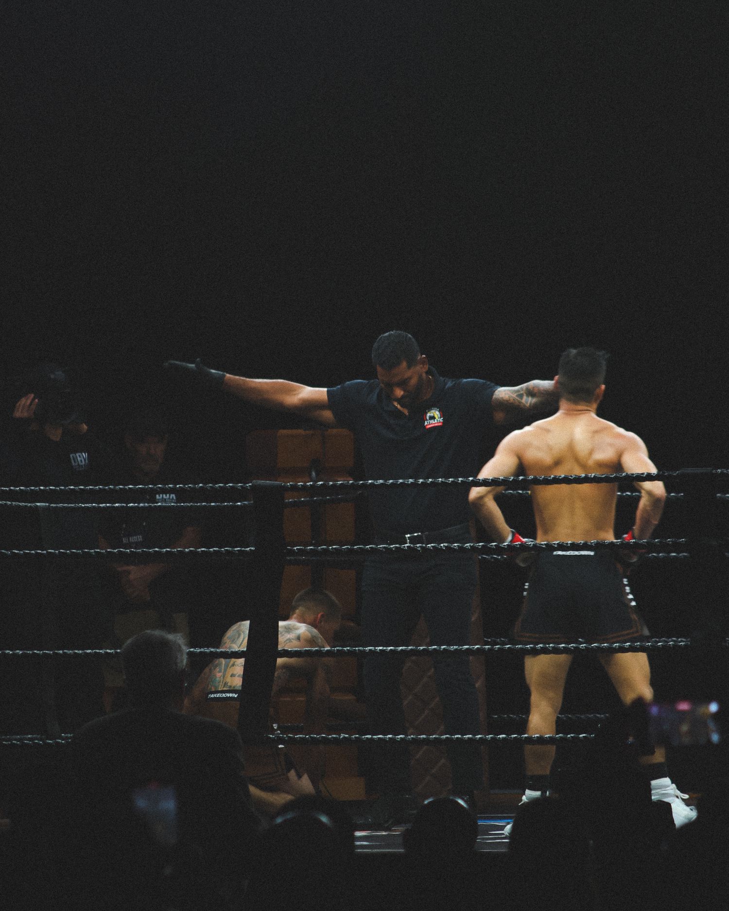 Wrestling referee stands between two competitors in a dimly lit ring during a match.