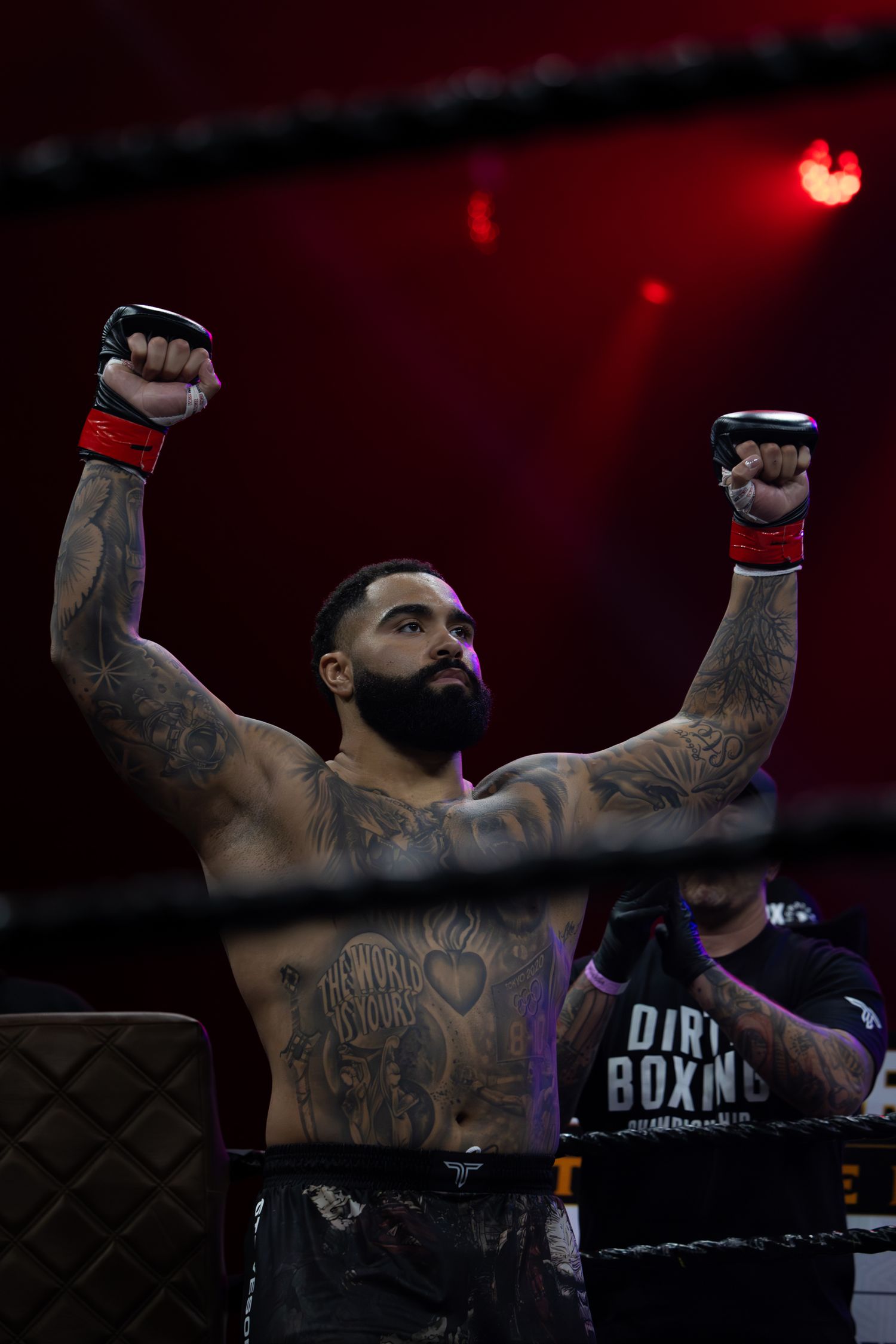 Tattooed performer raises arms triumphantly on stage under red lighting.