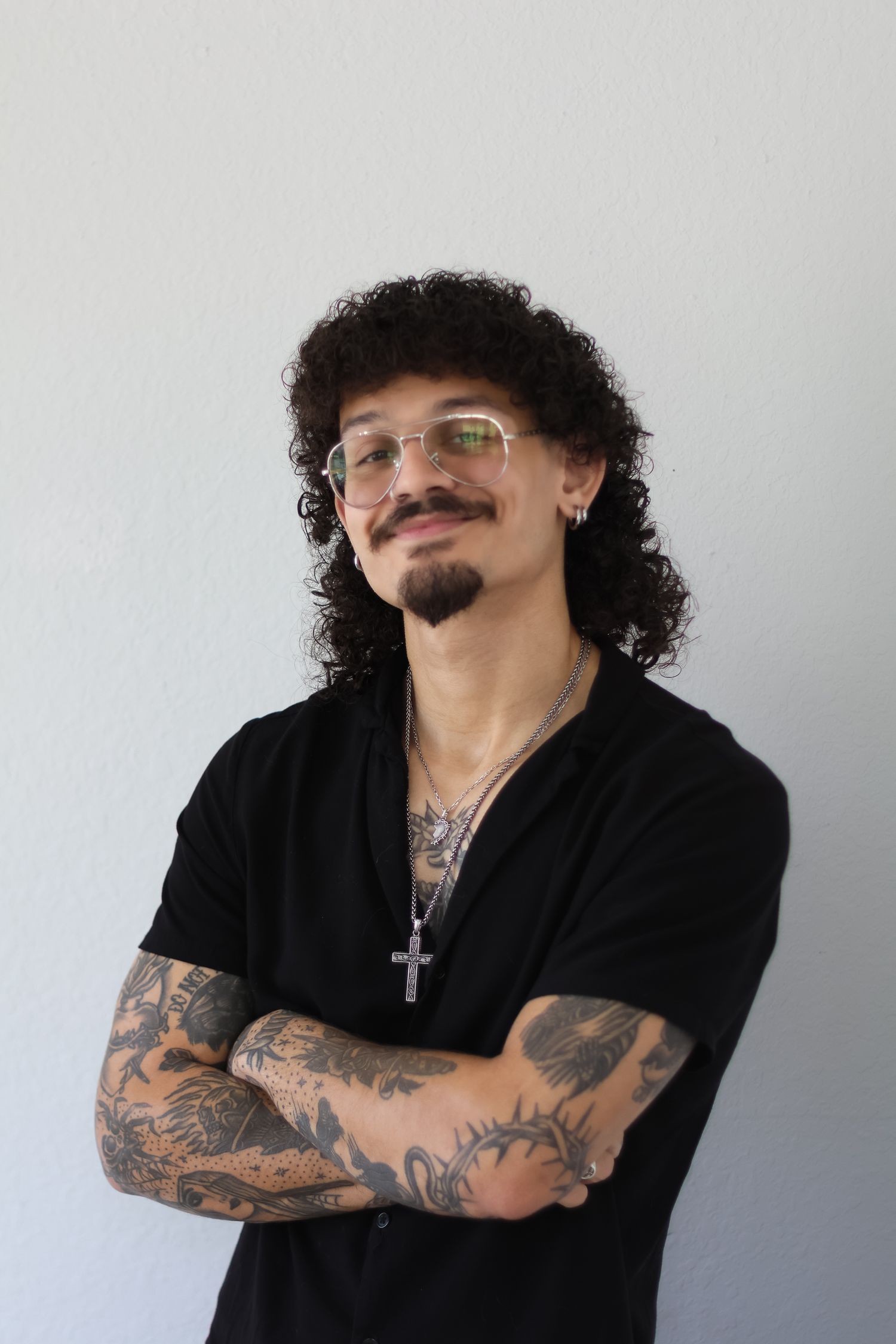 A person with curly hair and tattoos crosses their arms while wearing glasses and a black shirt.