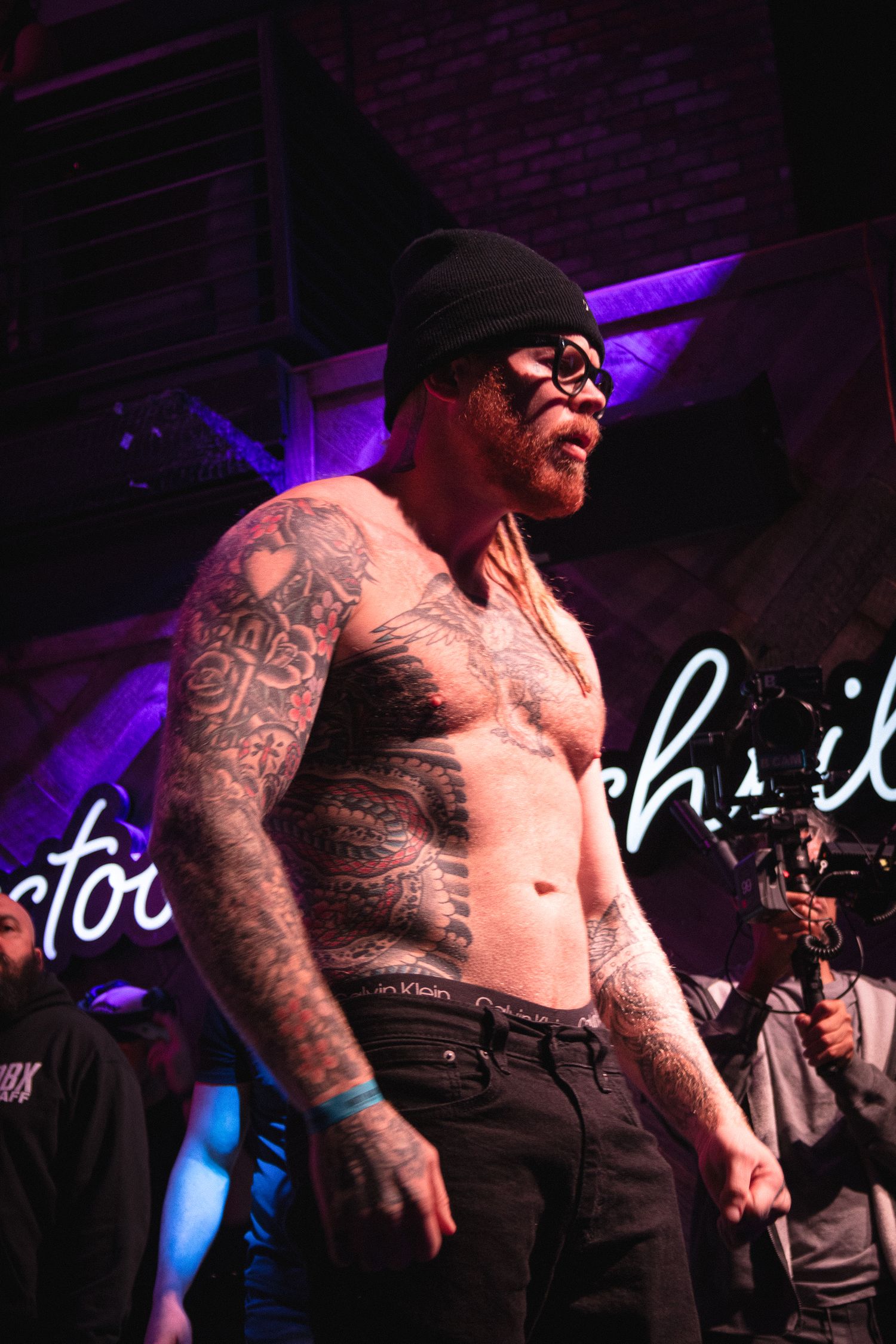 Tattooed performer wearing beanie and glasses performs shirtless under purple stage lights.