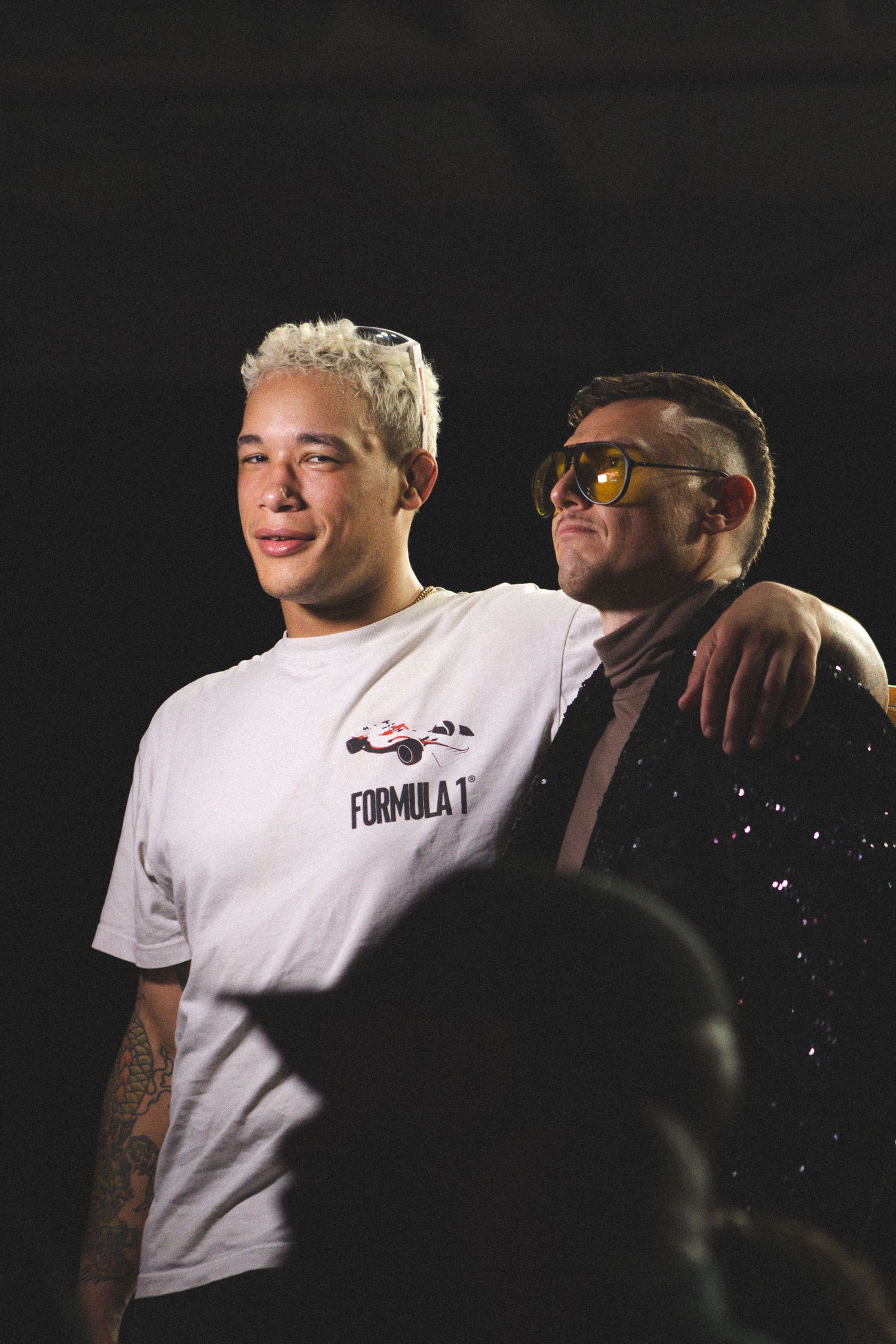 Two performers in sunglasses pose together, one wearing a white Formula 1 t-shirt.