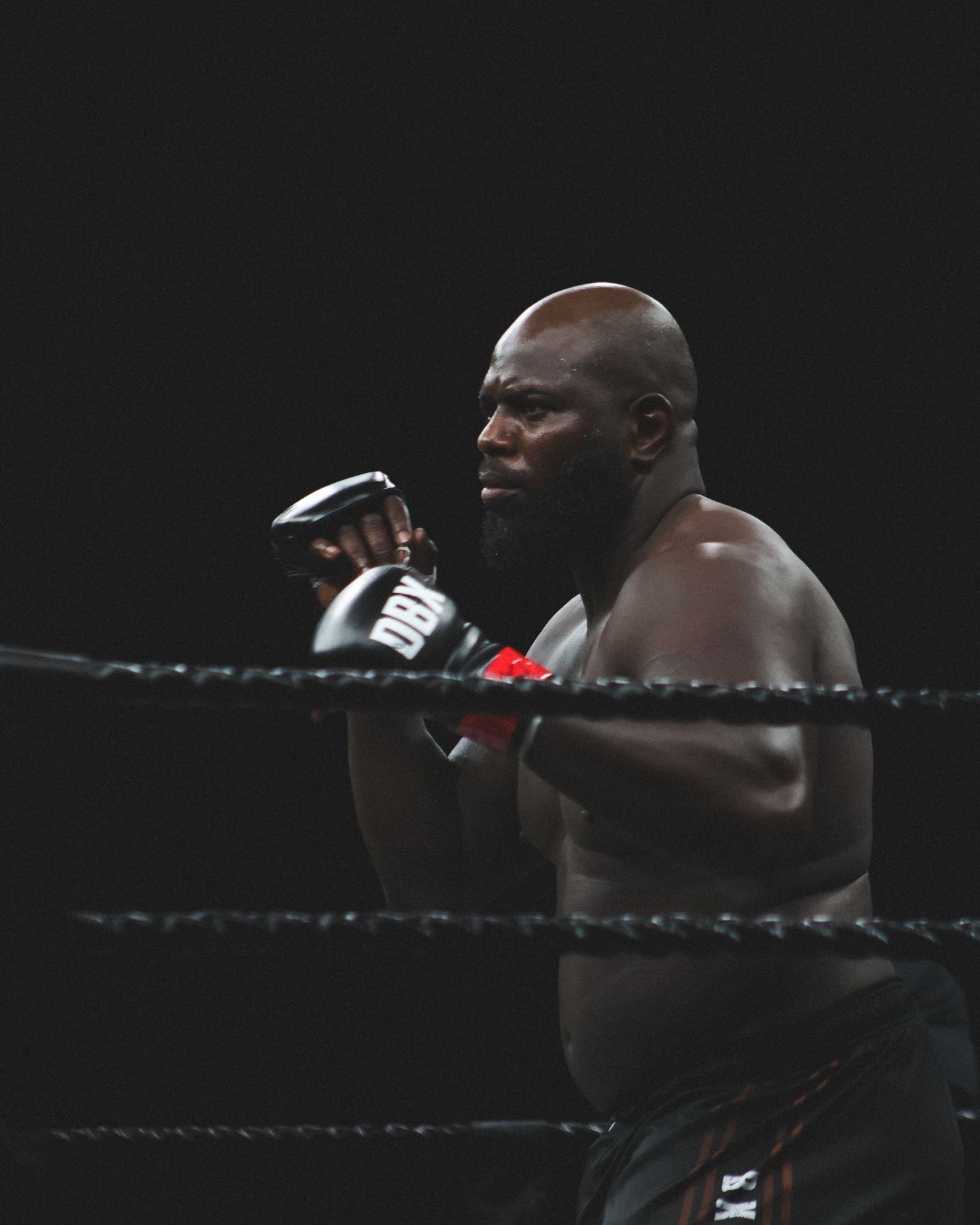 Muscular boxer in fighting stance viewed through ring ropes against dark background.
Rozenstruik