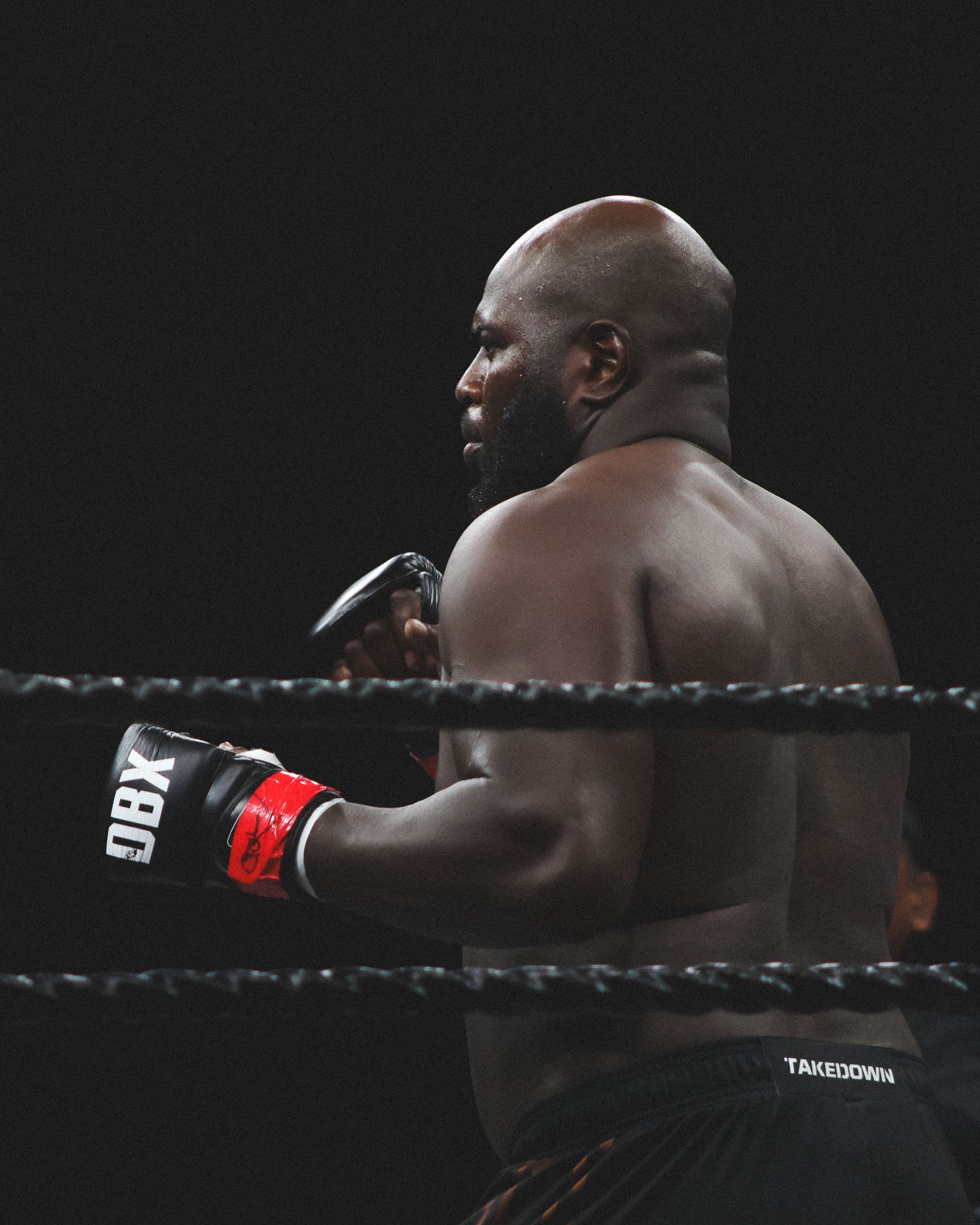 Side profile of a muscular boxer in black shorts positioned against the ring ropes.