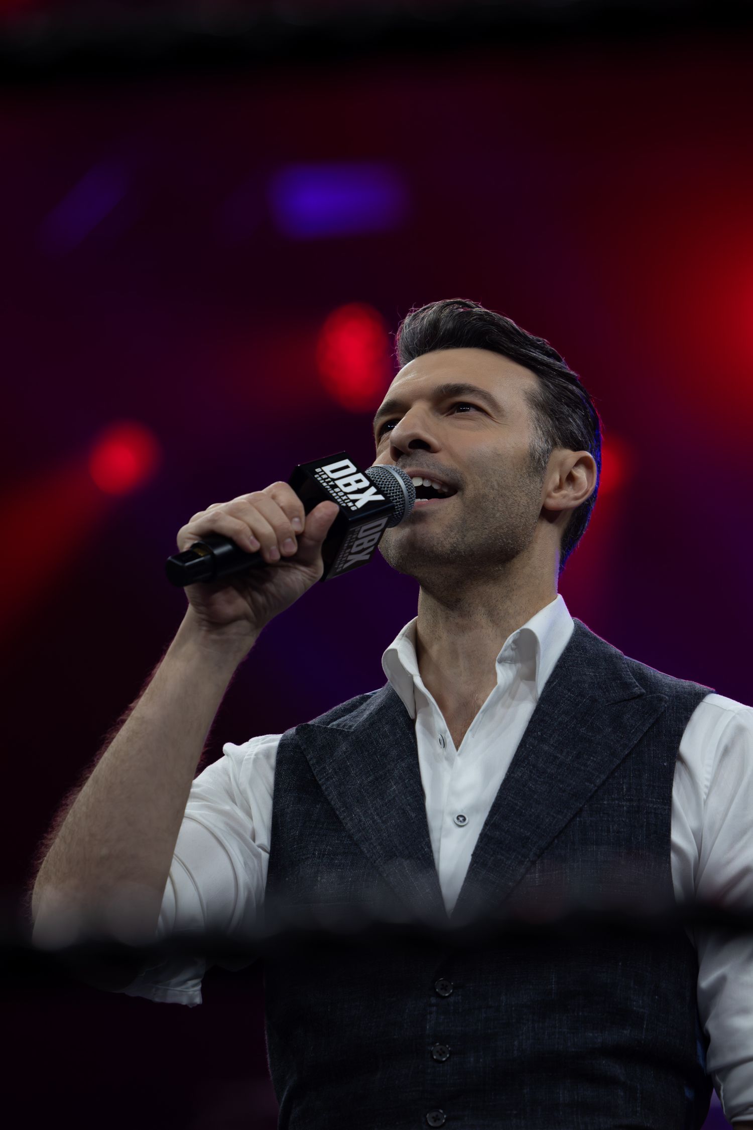 Singer in vest and white shirt performs emotionally under dramatic lighting.