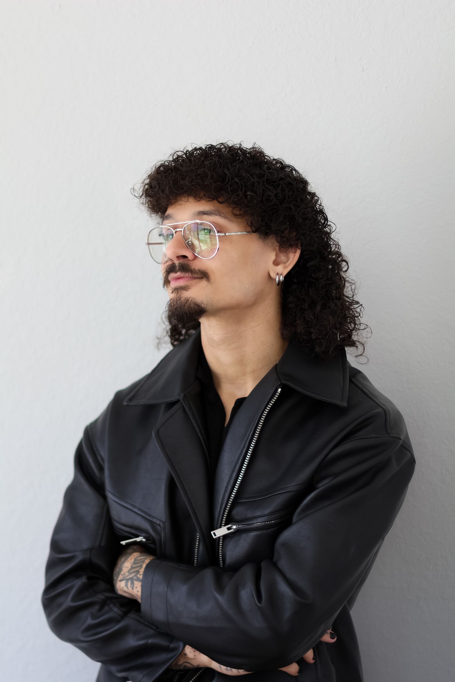 A person in a black leather jacket poses with crossed arms against a gray background.
